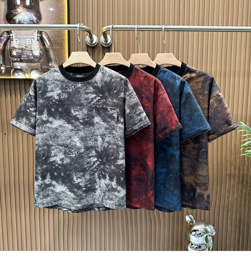 A row of four Common Theory Cloud Wash Tees hanging on wooden hangers from a silver clothing rack against a fluted wood wall paneling background. The foreground t-shirt features a black mineral wash texture with a dark ribbed crew neckline and a small patch pocket on the left chest. Behind it, the other three shirts display the same mineral wash pattern in red, blue, and brown color variants.
