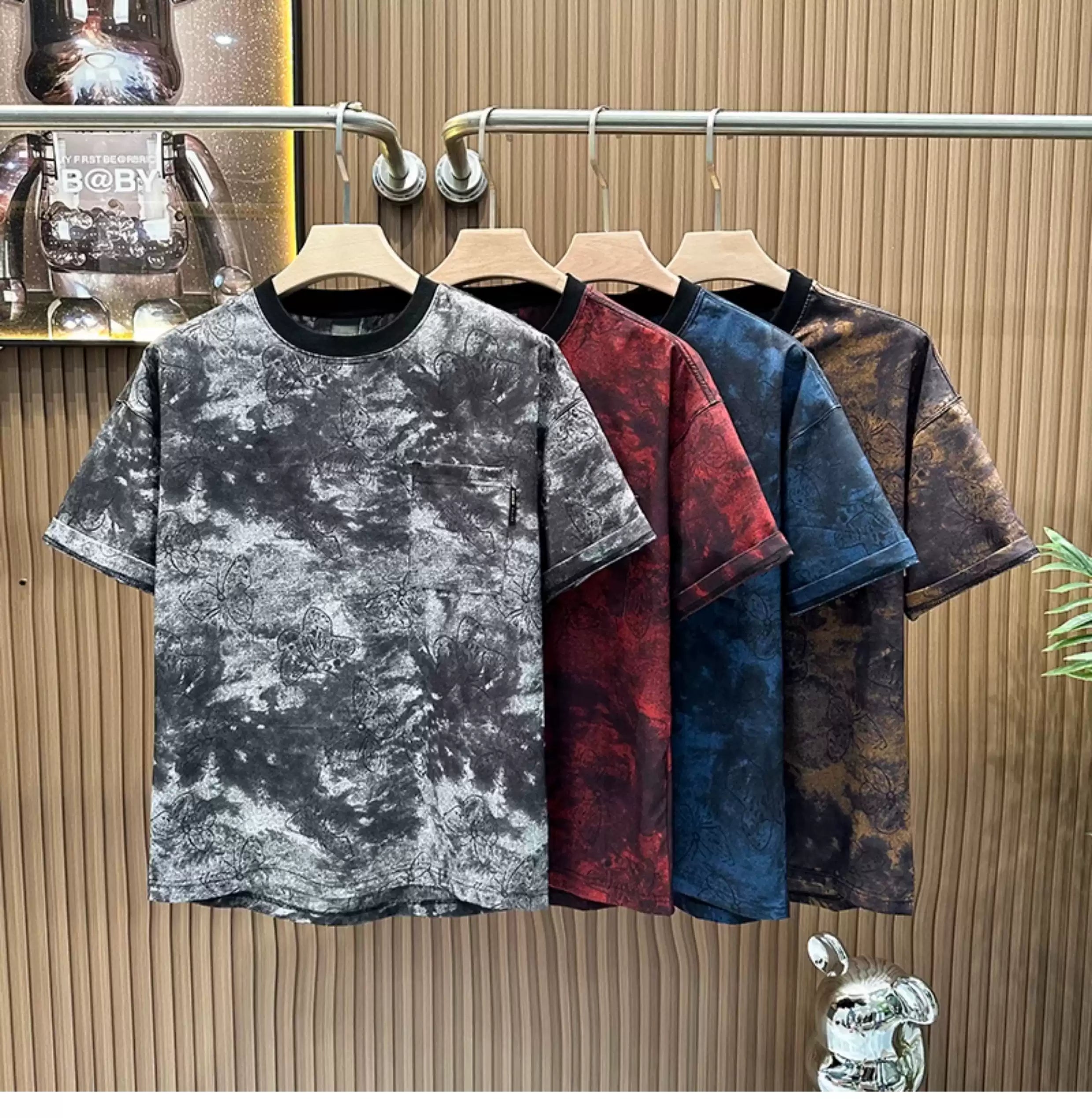 A row of four Common Theory Cloud Wash Tees hanging on wooden hangers from a silver clothing rack against a fluted wood wall paneling background. The foreground t-shirt features a black mineral wash texture with a dark ribbed crew neckline and a small patch pocket on the left chest. Behind it, the other three shirts display the same mineral wash pattern in red, blue, and brown color variants.