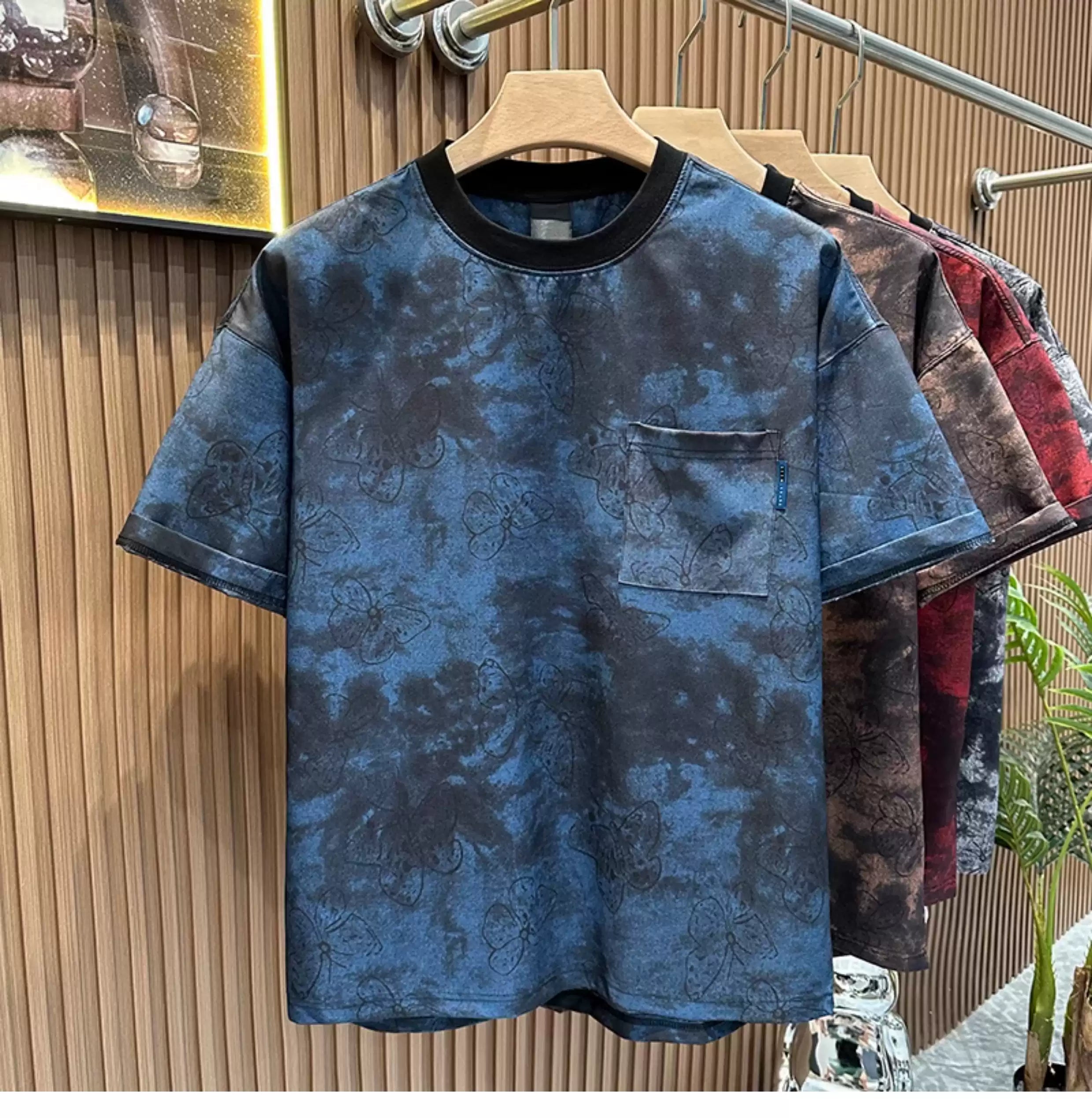Front view of the Common Theory Cloud Wash Tee in blue hanging on a wooden hanger, featuring a relaxed oversized fit, dropped shoulders, a dark ribbed crew neckline, and a functional patch pocket on the left chest with an all-over dark butterfly print over a mineral wash pattern. Other color variants (red, brown) are visible hanging behind it against a fluted wood wall paneling background.