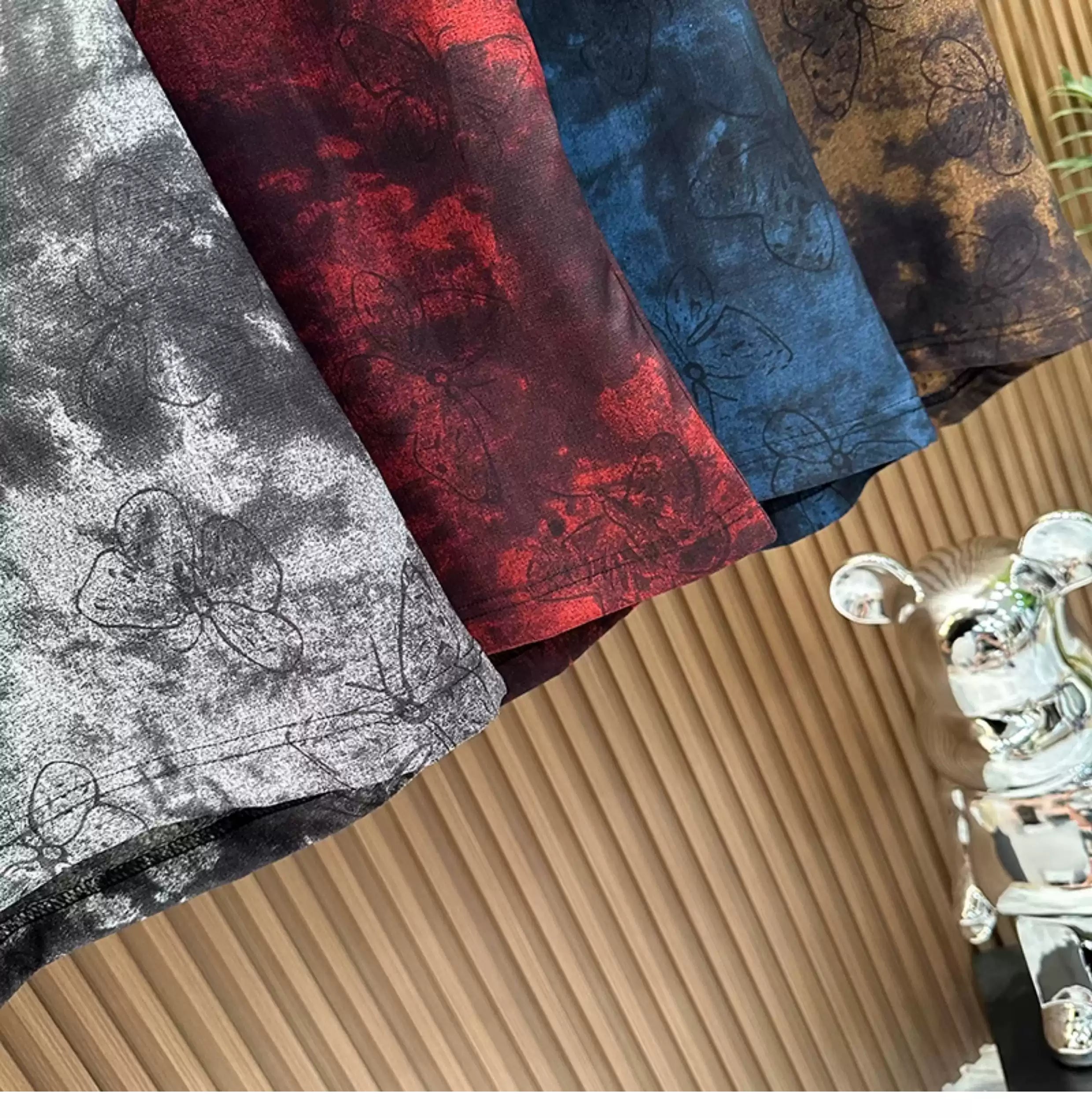 An angled, close-up view of four folded Common Theory Cloud Wash Tees stacked diagonally. The image highlights the detailed hemlines and the dark butterfly print overlaying the grey, crimson red, blue, and brown mineral wash textures against a fluted wood background.