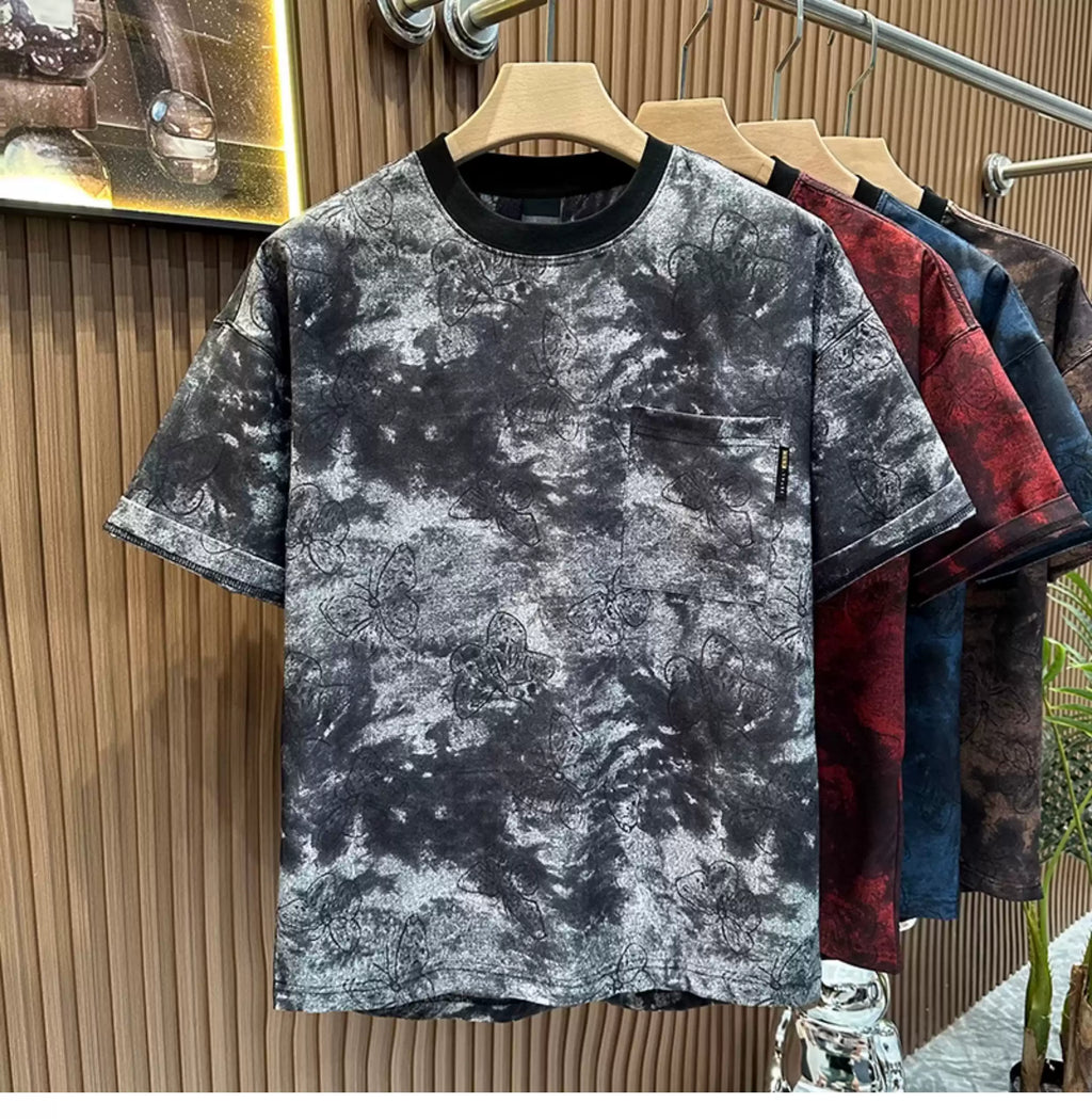 Front view of the Common Theory Cloud Wash Tee in a black and grey mineral wash pattern hanging on a wooden hanger, featuring an oversized relaxed fit, dropped shoulders, a solid black ribbed crew neckline, and a functional patch pocket on the left chest with an all-over dark butterfly print. Other color variants in red, blue, and brown are visible hanging behind it against a fluted wood wall paneling background.