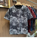 Front view of the Common Theory Cloud Wash Tee in a black and grey mineral wash pattern hanging on a wooden hanger, featuring an oversized relaxed fit, dropped shoulders, a solid black ribbed crew neckline, and a functional patch pocket on the left chest with an all-over dark butterfly print. Other color variants in red, blue, and brown are visible hanging behind it against a fluted wood wall paneling background.