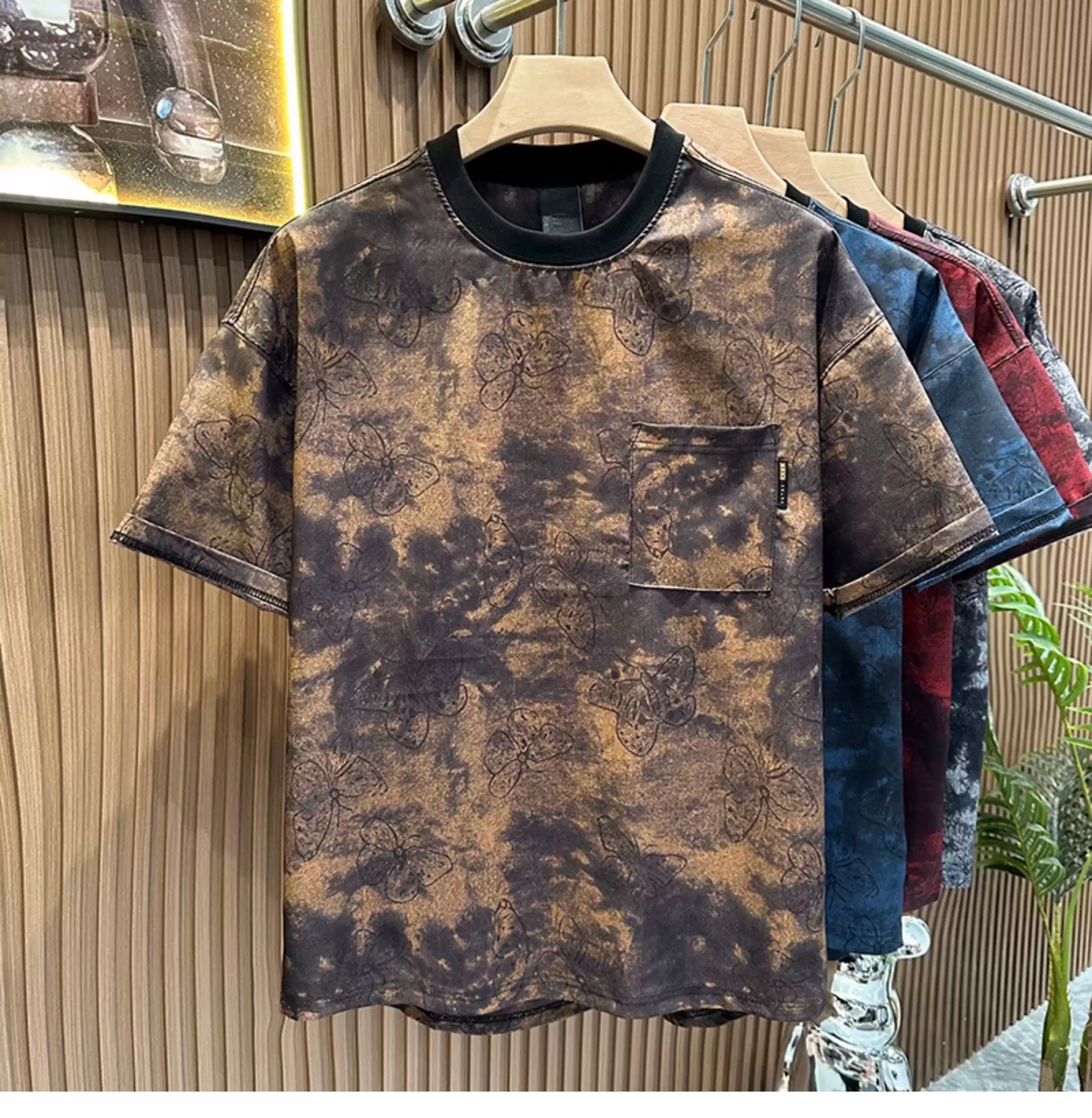 Front view of the Common Theory Cloud Wash Tee in vintage brown hanging on a wooden hanger, featuring a relaxed oversized fit, dropped shoulders, a solid black ribbed crew neckline, and a functional patch pocket on the left chest with an all-over dark butterfly print over a mineral wash pattern. Other color variants in blue and red are visible hanging behind it against a fluted wood wall paneling background.