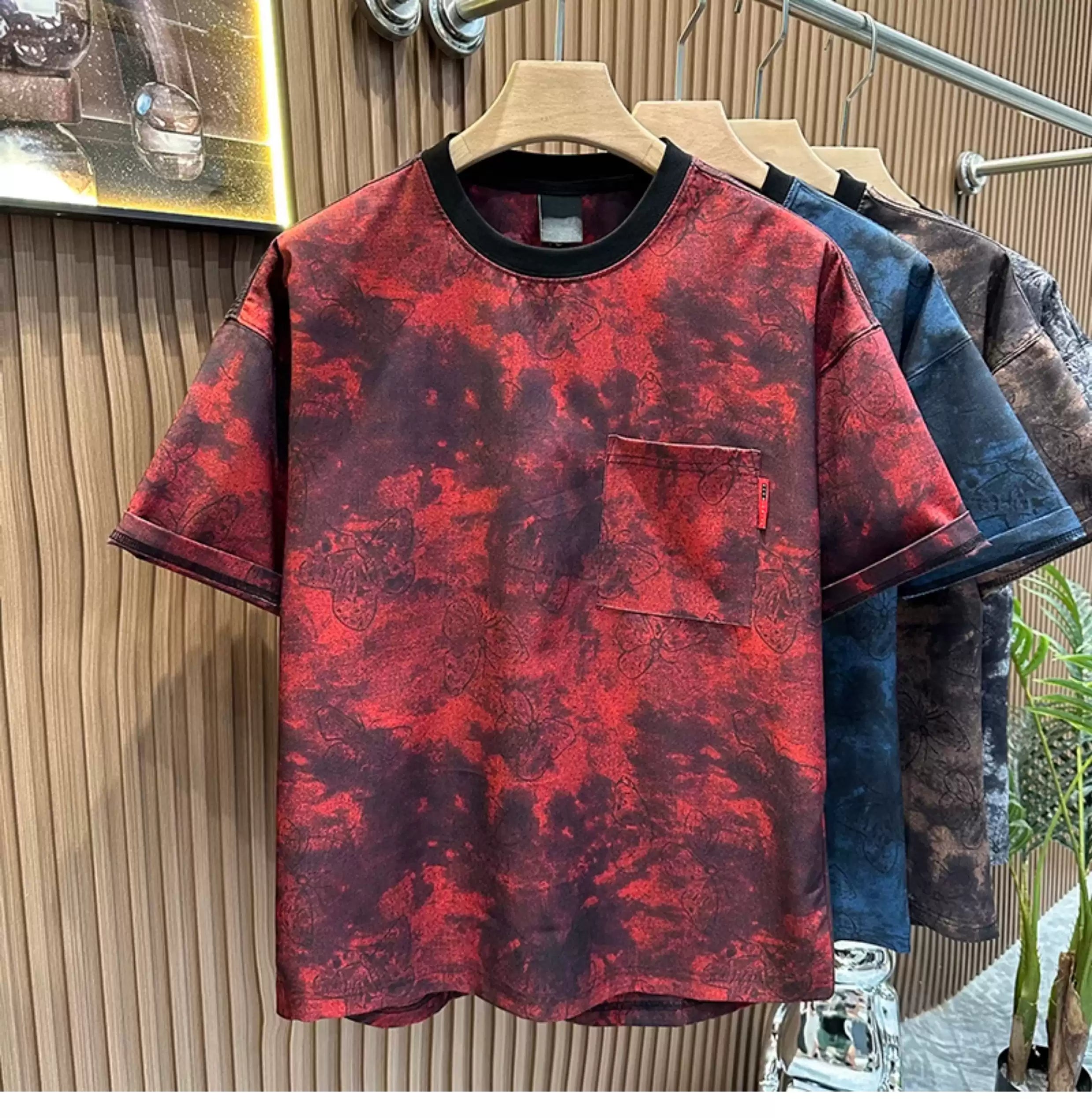 Front view of the Common Theory Cloud Wash Tee in crimson red hanging on a wooden hanger, featuring a relaxed oversized fit, dropped shoulders, a dark ribbed crew neckline, and a functional patch pocket on the left chest with an all-over dark butterfly print over a red and black mineral wash pattern. Other color variants in blue, black, and brown are visible hanging behind it against a fluted wood wall paneling background.