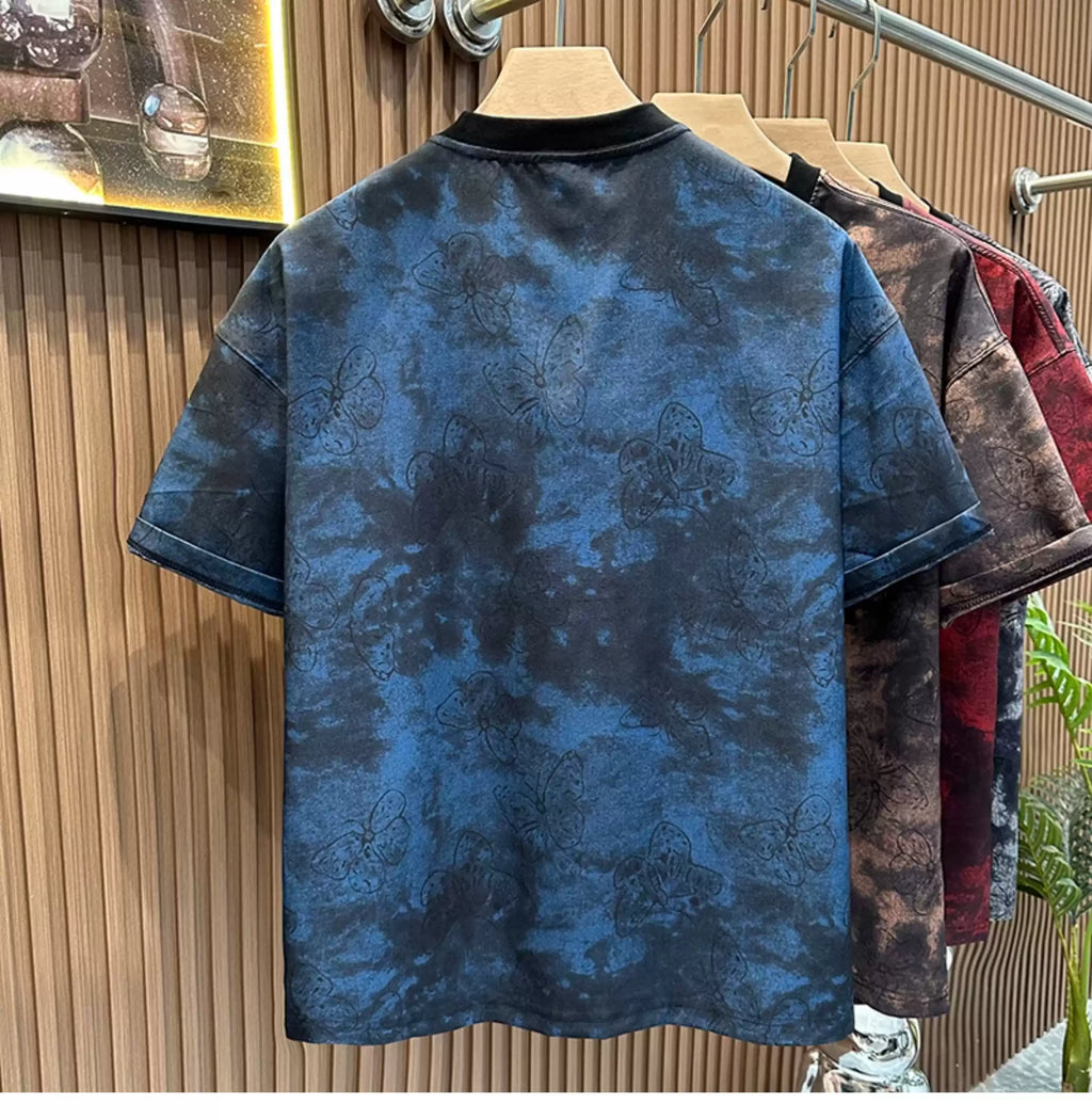 Rear view of the Common Theory Cloud Wash Tee in blue hanging on a wooden hanger, highlighting its oversized streetwear silhouette, dropped shoulders, and a solid black ribbed crew neckline. The fabric showcases an all-over dark butterfly pattern overlaid on a blue and black mineral wash texture, with other color variants (brown, red) hanging behind it against a fluted wood wall paneling background.