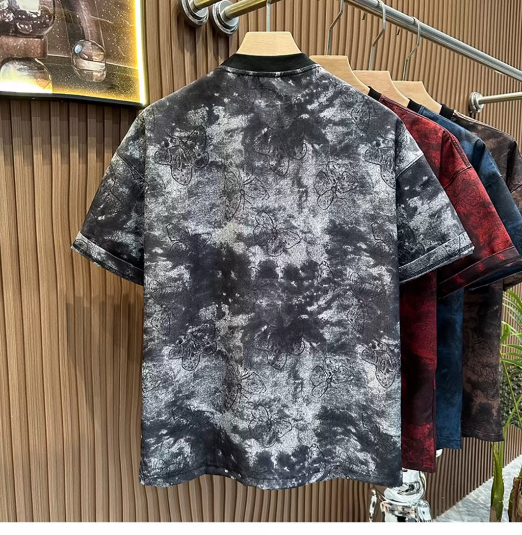 Rear view of the Common Theory Cloud Wash Tee in black hanging on a wooden hanger, highlighting its oversized streetwear silhouette, dropped shoulders, and a solid black ribbed crew neckline. The fabric showcases an all-over dark butterfly pattern overlaid on a grey and black mineral wash texture, with other color variants in crimson red, blue, and brown hanging behind it against a fluted wood wall paneling background.