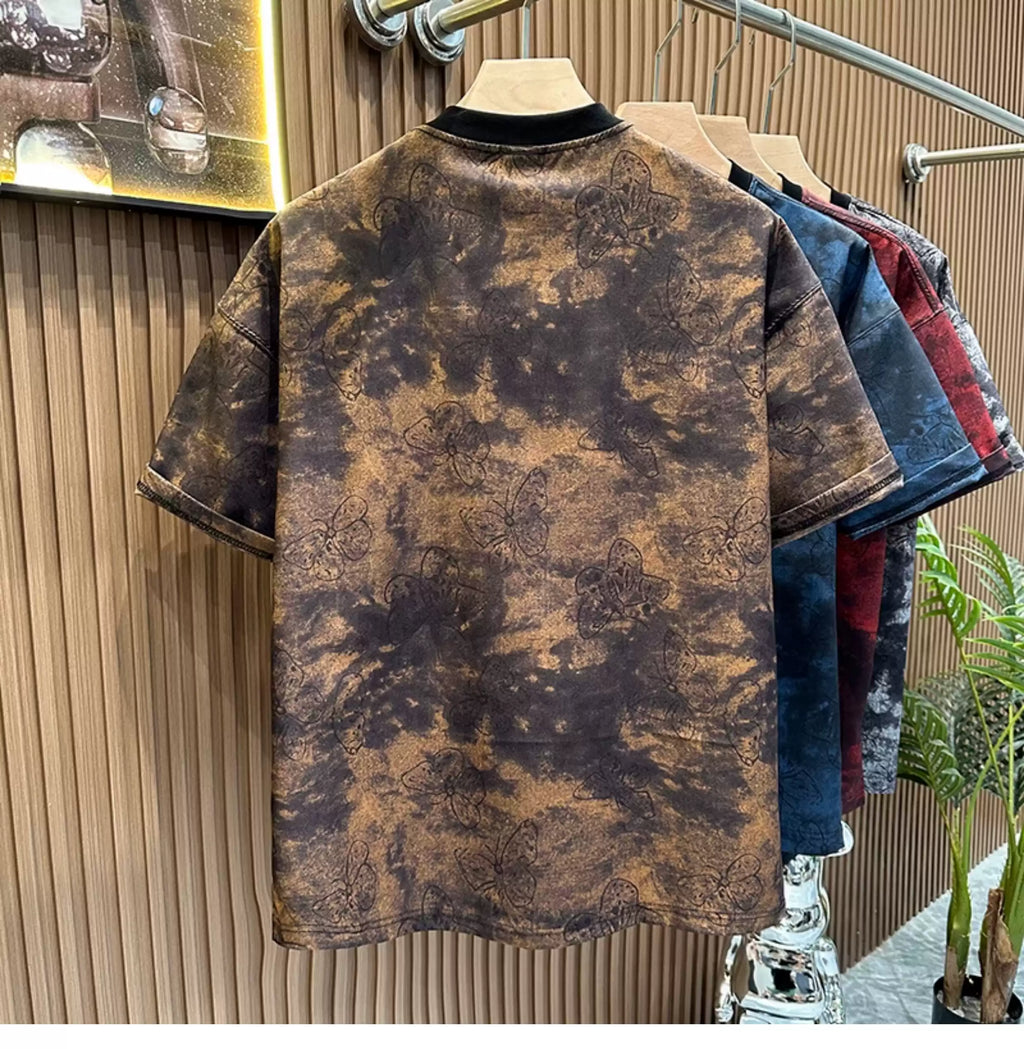 Rear view of the Common Theory Cloud Wash Tee in vintage brown hanging on a wooden hanger, highlighting its oversized streetwear silhouette, dropped shoulders, and a solid black ribbed crew neckline. The fabric showcases an all-over dark butterfly pattern overlaid on a brown and black mineral wash texture, with other color variants in blue, red, and black hanging behind it against a fluted wood wall paneling background.