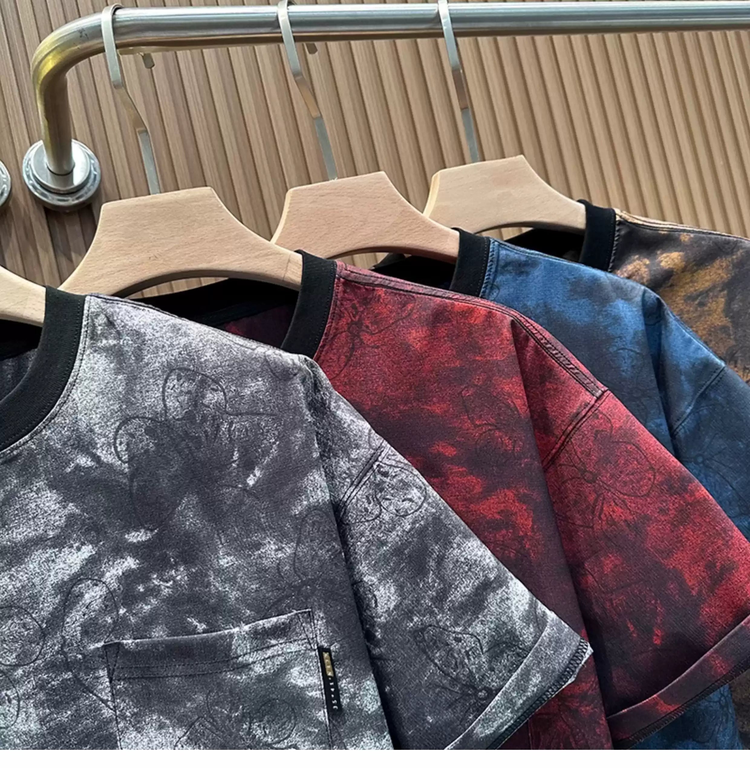 A close-up, angled view of four Common Theory Cloud Wash Tees hanging on wooden hangers from a silver clothing rack. The front shirt features a black and grey mineral wash pattern with an all-over dark butterfly print, a solid black ribbed crew neckline, and a small black brand tag on the edge of the left chest pocket. Hanging closely behind it are the crimson red, midnight blue, and vintage brown color variants, showcasing their matching mineral wash textures and dark necklines against a fluted wood wall p