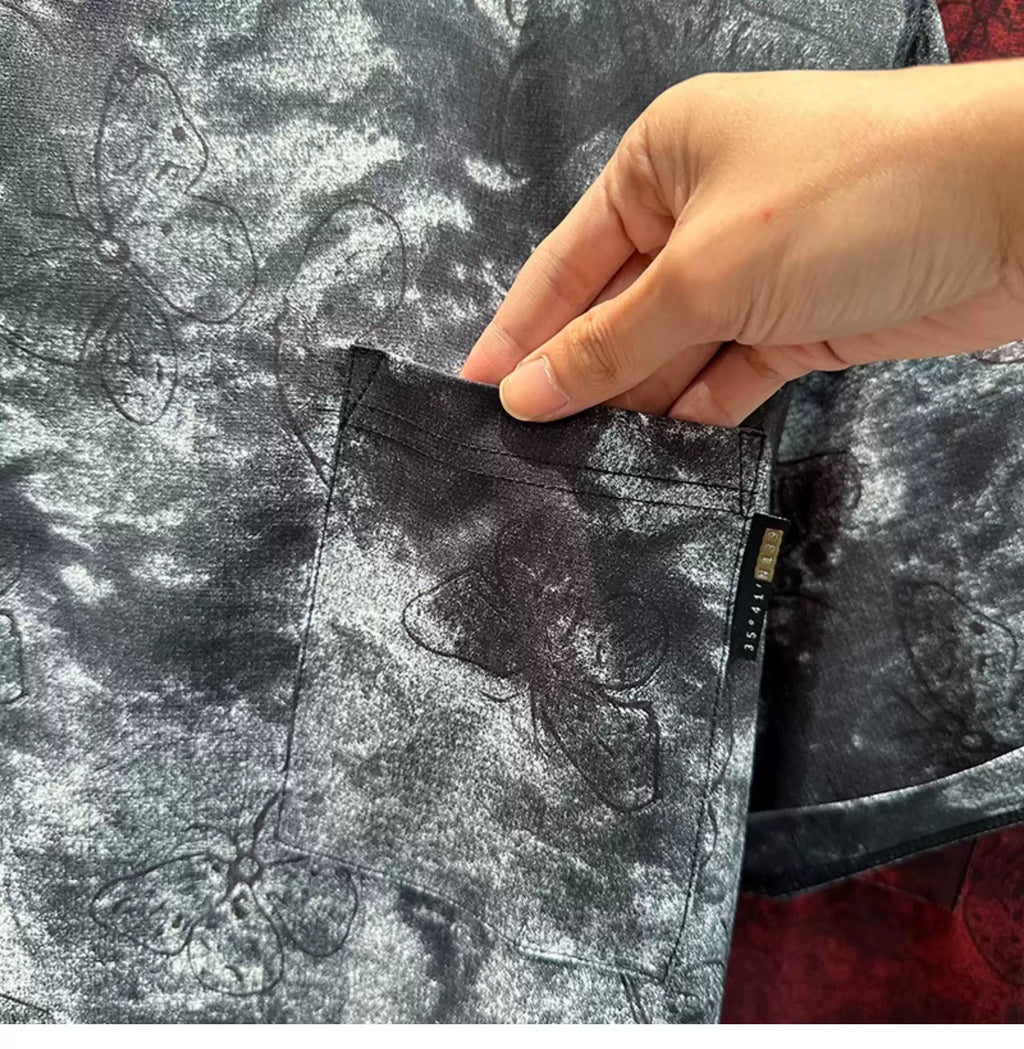 Close-up view of a hand lifting the chest pocket on the Common Theory Cloud Wash Tee, showcasing the grey and black mineral wash pattern, embroidered butterfly detail, and a small black brand tag.