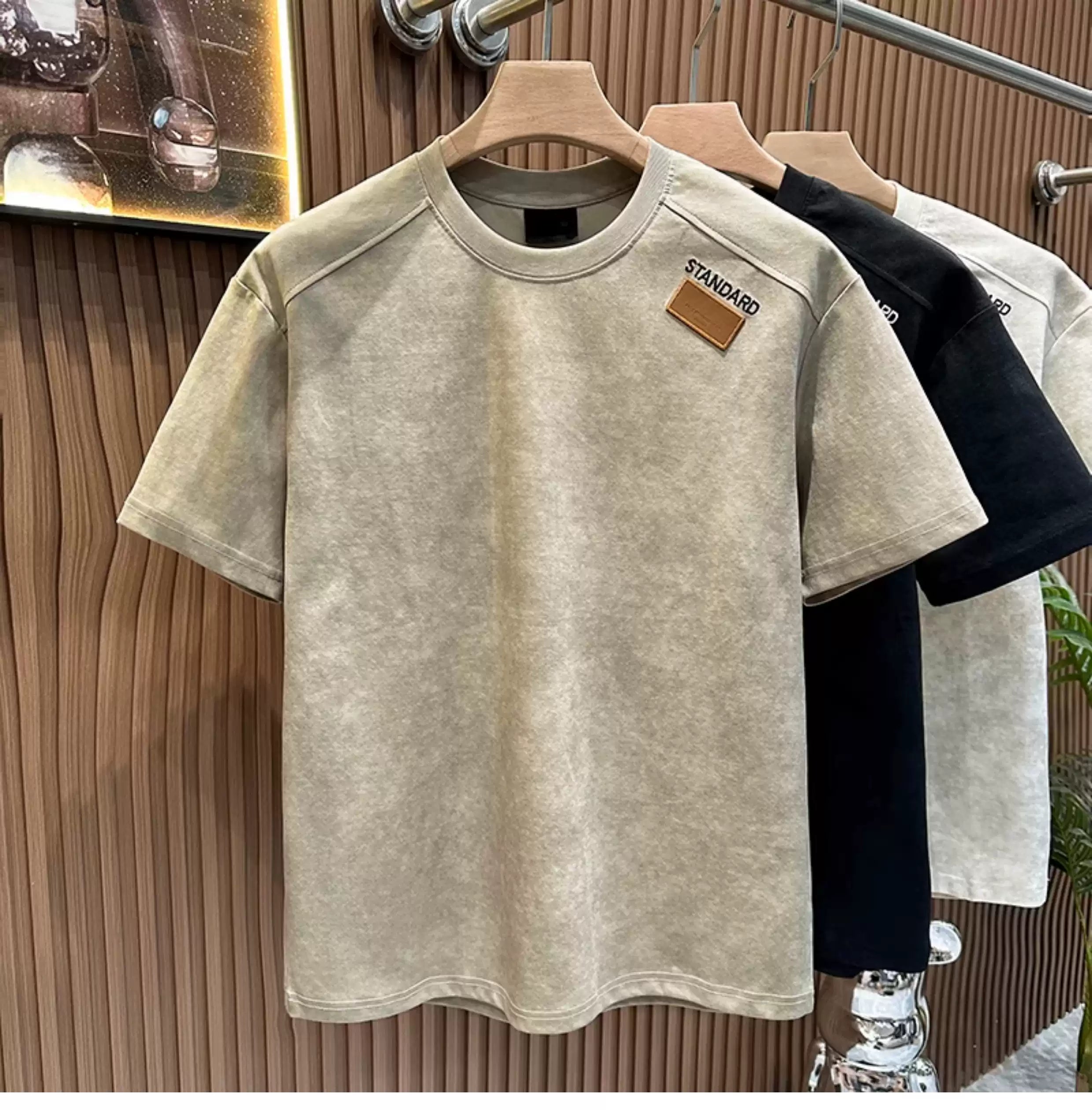 Front view of the Common Theory Embroidered Heavyweight Cotton T-Shirt in stone gray hanging on a wooden hanger, featuring a relaxed fit, ribbed crew neckline, and a small brown square "STANDARD" patch on the left chest, with black and white color variants visible hanging in the background against a fluted wood wall paneling.
