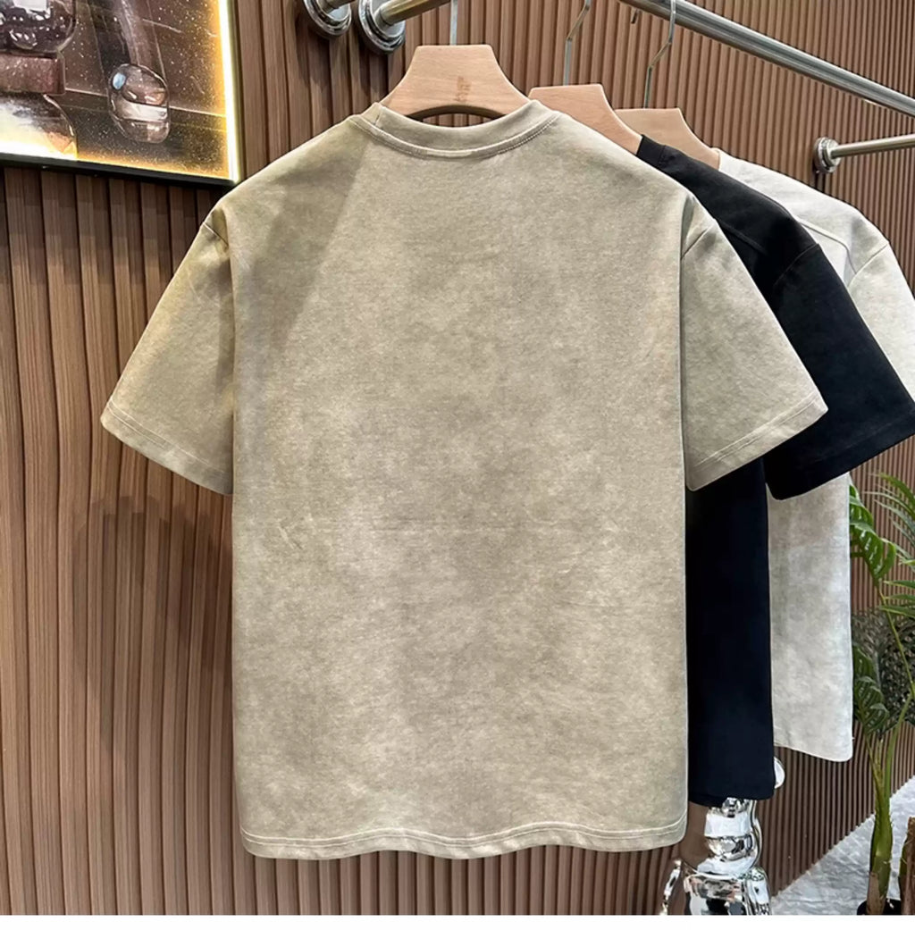 Rear view of the Common Theory Embroidered Heavyweight Cotton T-Shirt in stone gray hanging on a wooden hanger, displaying its clean finish and relaxed silhouette, with black and white color variants visible hanging in the background against a fluted wood wall paneling.