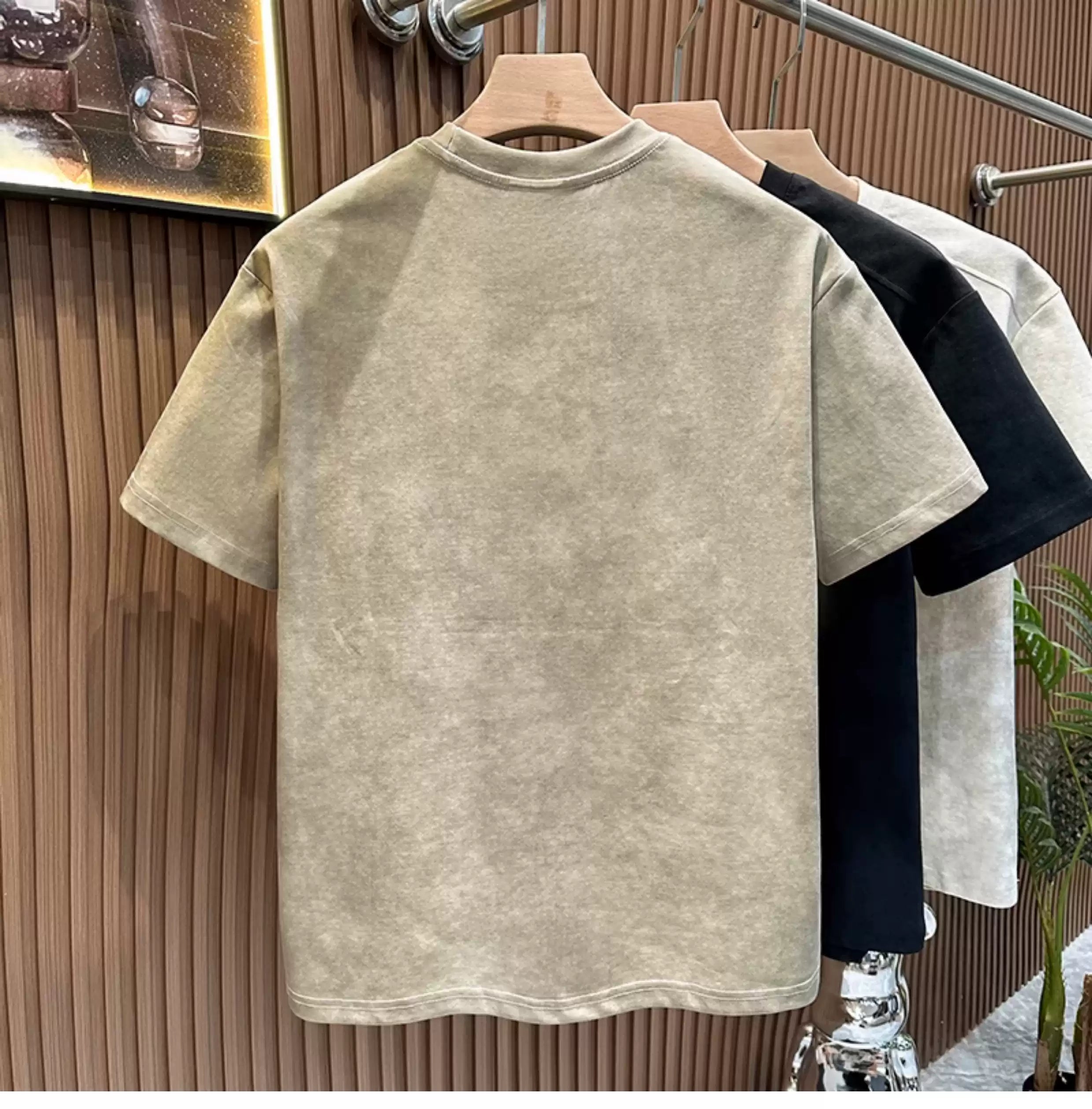 Rear view of the Common Theory Embroidered Heavyweight Cotton T-Shirt in stone gray hanging on a wooden hanger, displaying its clean finish and relaxed silhouette, with black and white color variants visible hanging in the background against a fluted wood wall paneling.
