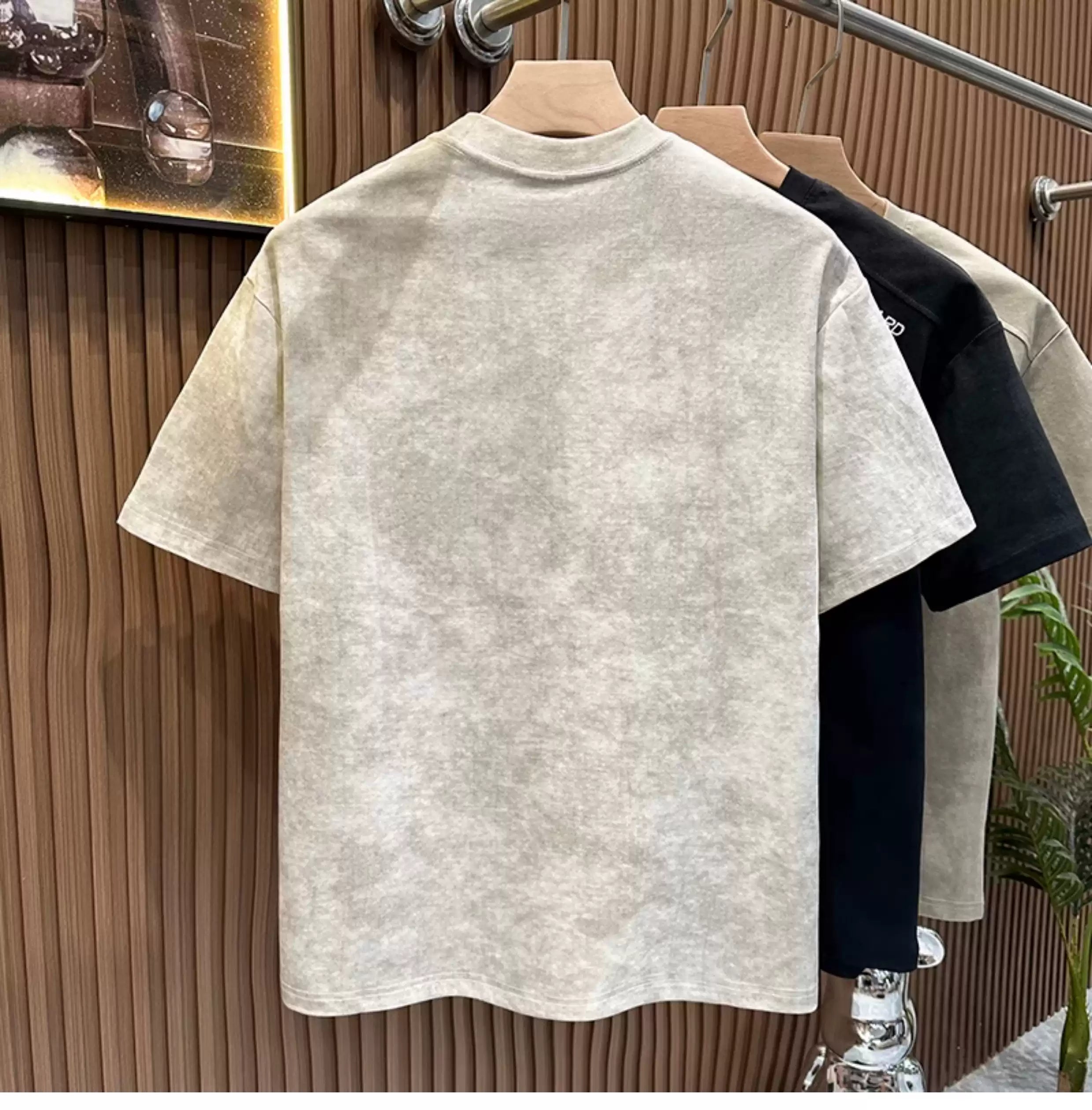 Rear view of the Common Theory Embroidered Heavyweight Cotton T-Shirt in stone gray hanging on a wooden hanger, highlighting its clean stitching, relaxed fit, and thick collar band, with black and white color variants visible on a clothing rack against a fluted wood accent wall.
