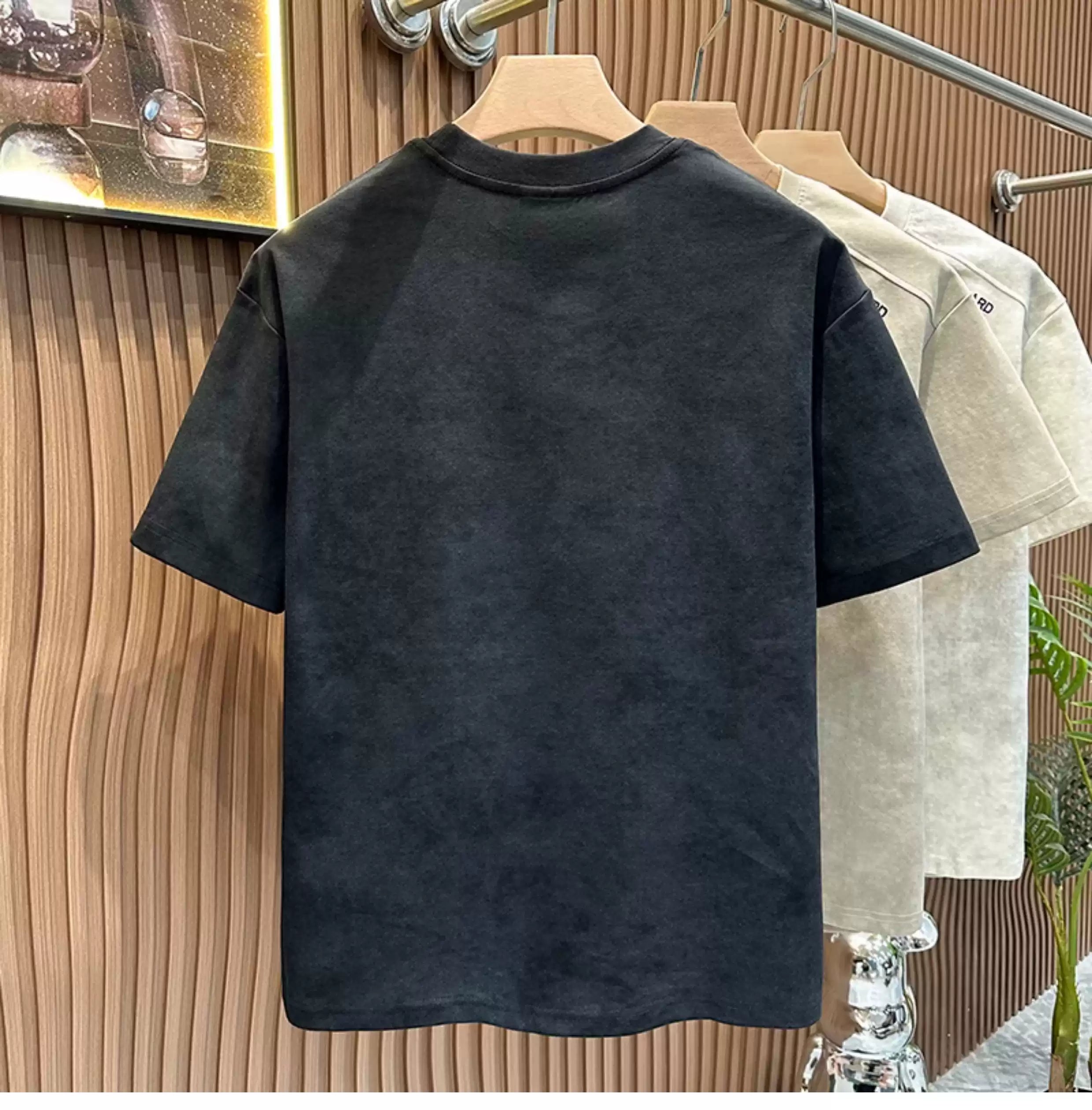 Front view of the Common Theory Embroidered Heavyweight Cotton T-Shirt in black hanging on a wooden hanger, highlighting its ribbed crew neckline and the small brown square "STANDARD" patch on the left chest, with stone gray and white variants visible in the background against a fluted wood wall paneling.