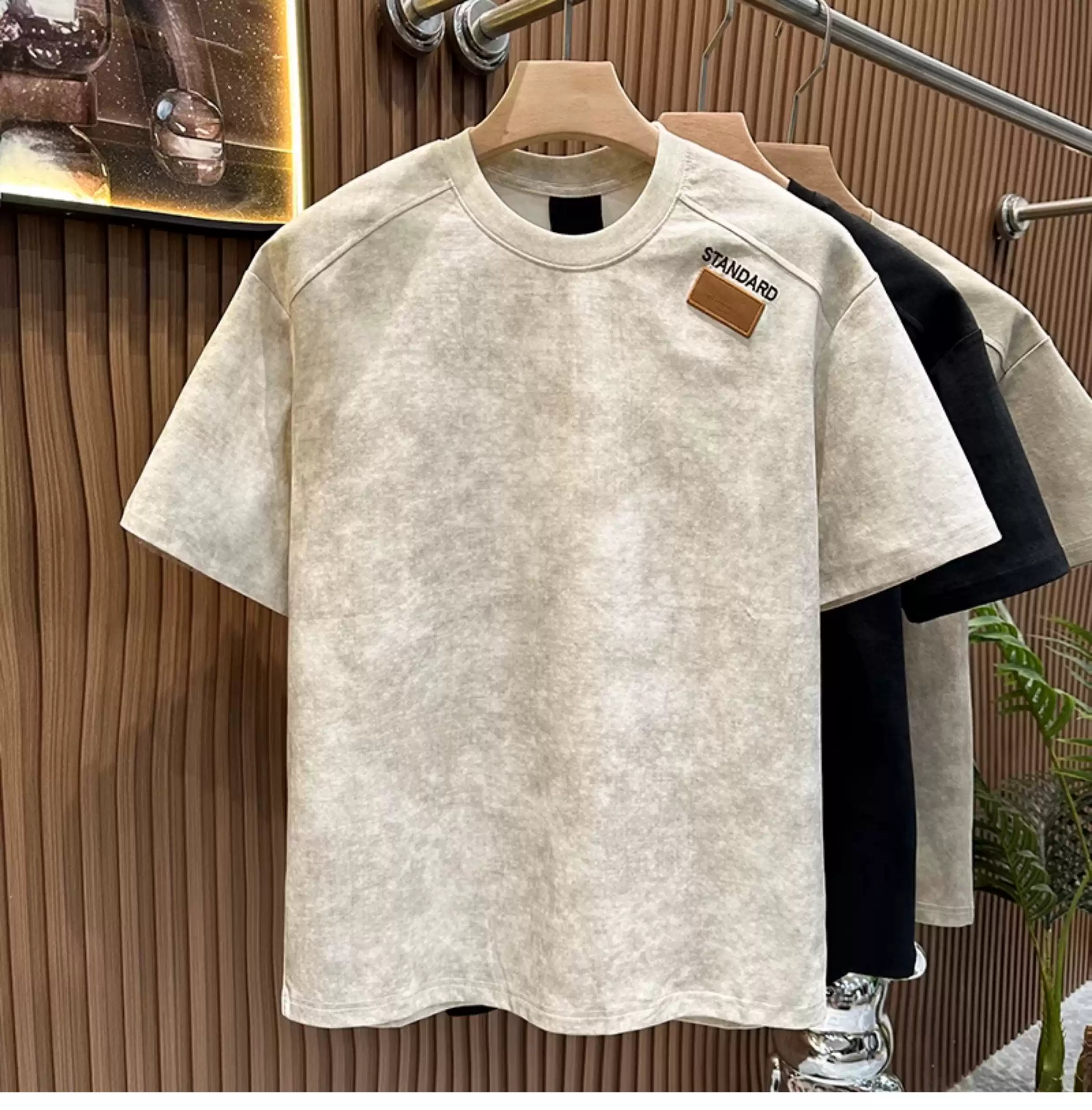 Front view of the Common Theory Embroidered Heavyweight Cotton T-Shirt in sand hanging on a wooden hanger, featuring a relaxed fit, a ribbed crew neckline, and a small brown square "STANDARD" patch on the left chest, with black and stone gray color variants visible hanging in the background against a fluted wood wall paneling.