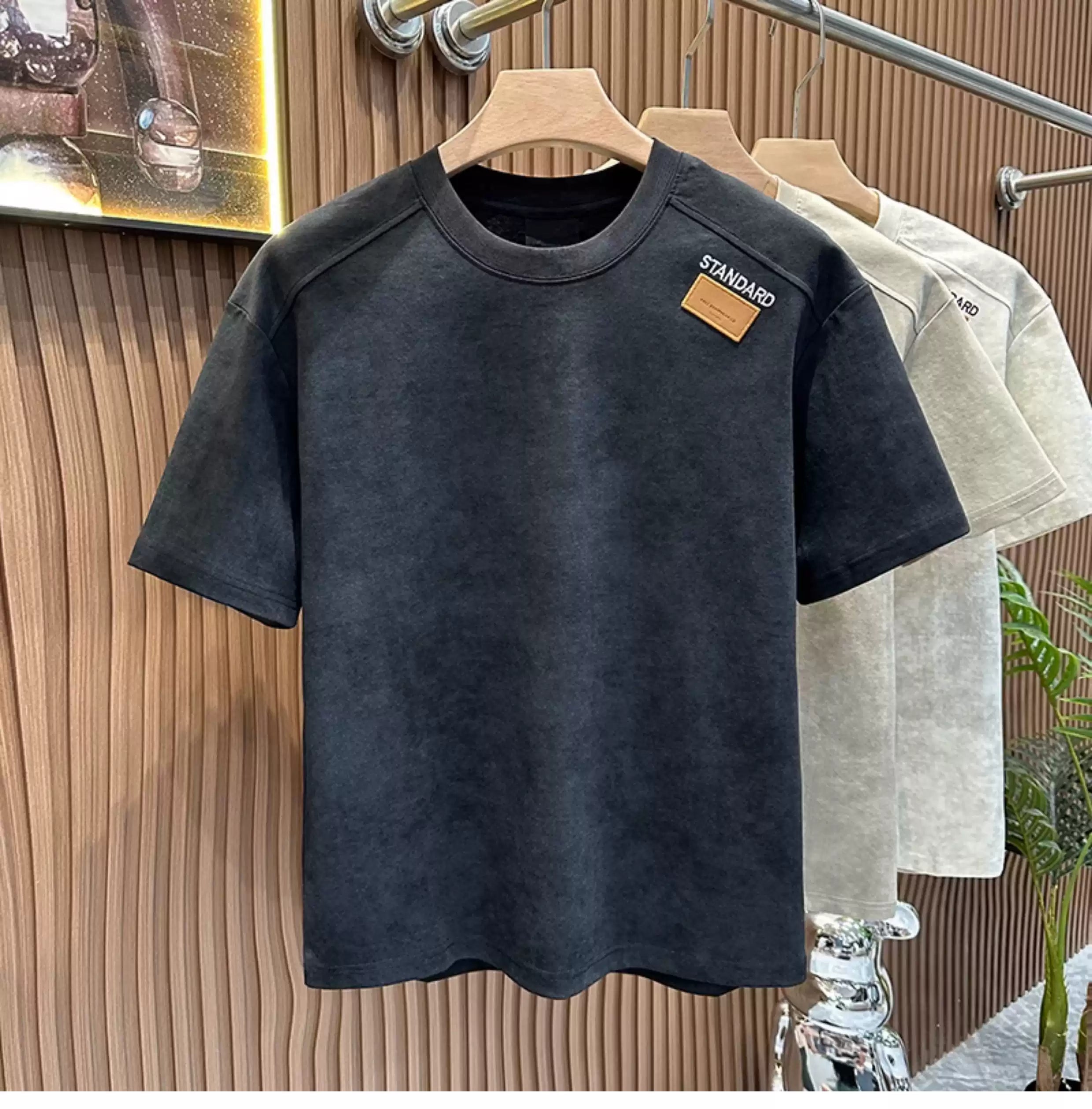 Front view of the Common Theory Embroidered Heavyweight Cotton T-Shirt in black with an acid-wash texture hanging on a wooden hanger, featuring a relaxed fit, a ribbed crew neckline, and a small brown square "STANDARD" patch on the left chest, with stone gray and sand variants visible hanging in the background against a fluted wood wall paneling