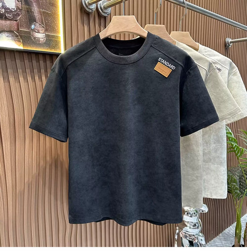 Front view of the Common Theory Embroidered Heavyweight Cotton T-Shirt in black with an acid-wash texture hanging on a wooden hanger, featuring a relaxed fit, a ribbed crew neckline, and a small brown square "STANDARD" patch on the left chest, with stone gray and sand variants visible hanging in the background against a fluted wood wall paneling