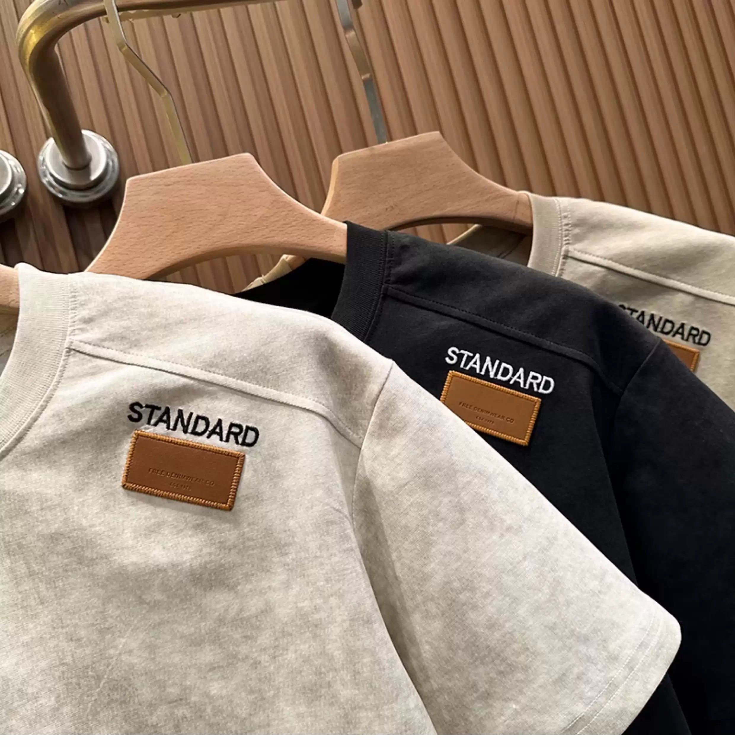 A close-up view of three Common Theory Embroidered Heavyweight Cotton T-Shirts hanging side-by-side on wooden hangers, showing the stone gray, black, and sand color variants. The focus highlights their ribbed crew necklines, relaxed fits, and the signature small brown square "STANDARD" patch on each left chest against a fluted wood wall paneling background.