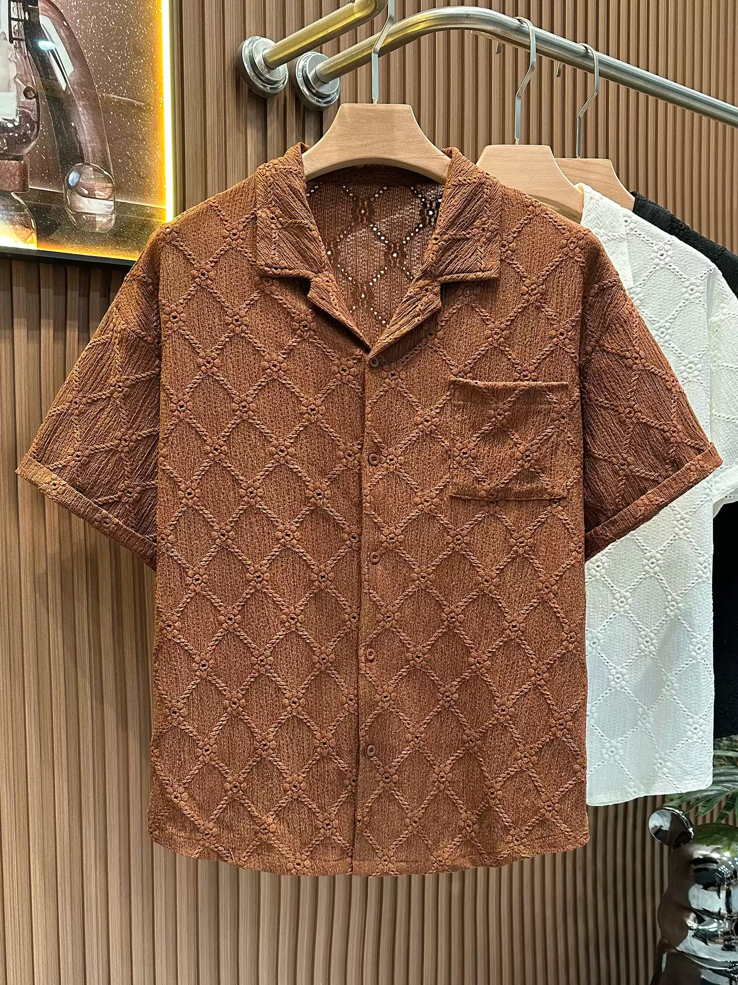 The Common Theory Riviera Cocoa Brown Resort Shirt hanging on a wooden clothes hanger, showcasing its relaxed camp collar, front chest pocket, and intricate geometric diamond open-knit pattern against a fluted wood wall paneling background.