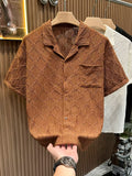 Front view of the Common Theory Riviera Cocoa Brown Resort Shirt on a wooden hanger, featuring a distinctive geometric diamond and floral open-knit pattern, a relaxed camp collar, a front chest pocket, and short sleeves, with a person's hand visible at the bottom hem.
