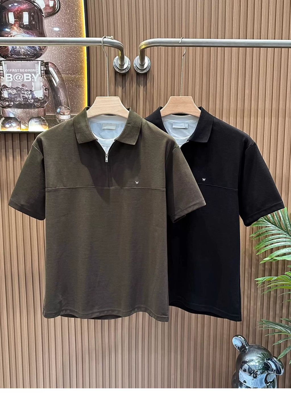 Front view of the Common Theory Signature Quarter-Zip Polo in dark olive hanging on a wooden hanger, featuring a structured silhouette, a mid-chest horizontal seam, a silver zipper, a ribbed collar, and a small embroidered chest logo. A black color variant is visible hanging directly behind it against a fluted wood wall paneling background.