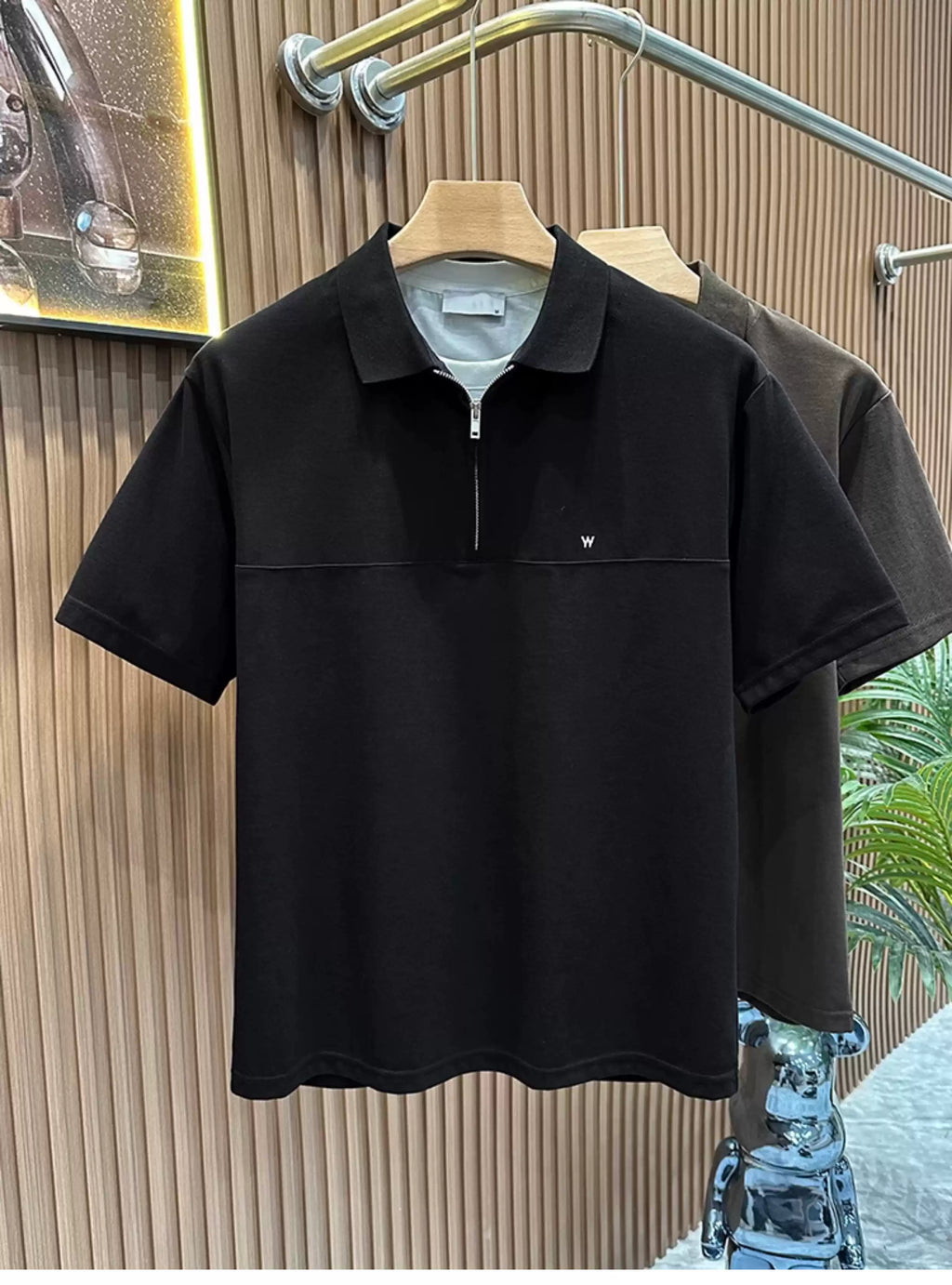 Front view of the Common Theory Signature Quarter-Zip Polo in black hanging on a wooden hanger, featuring a structured silhouette, a mid-chest horizontal seam, a silver zipper, a ribbed collar, and a small minimalist embroidered logo on the left chest. A dark olive color variant is visible hanging directly behind it against a fluted wood wall paneling background.