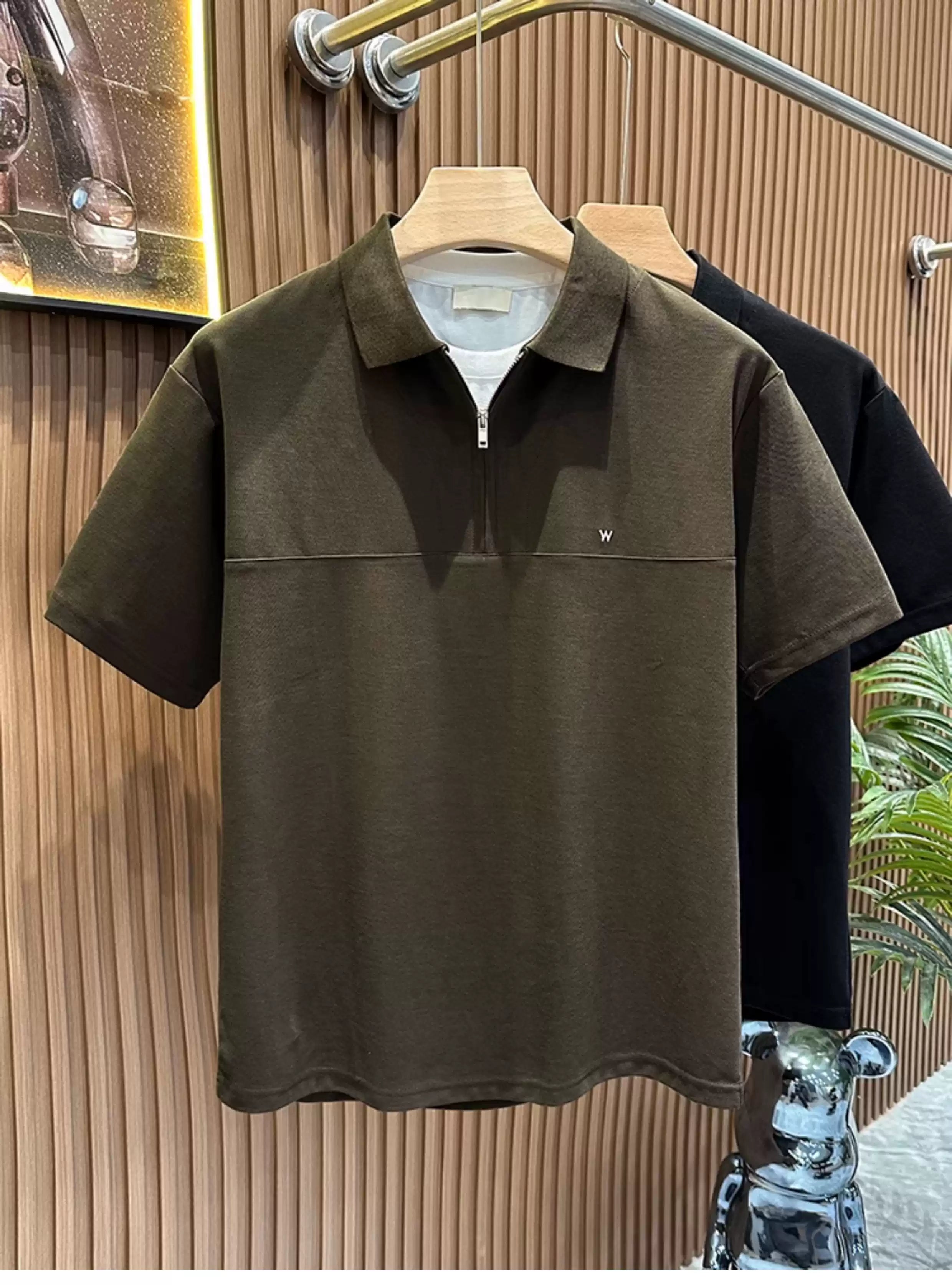 Front view of the Common Theory Signature Quarter-Zip Polo in dark olive hanging on a wooden hanger, highlighting its classic ribbed collar, silver quarter-zip closure, structured silhouette, and a small minimalist embroidered logo on the left chest. A black color variant is visible hanging directly behind it against a fluted wood wall paneling background.
