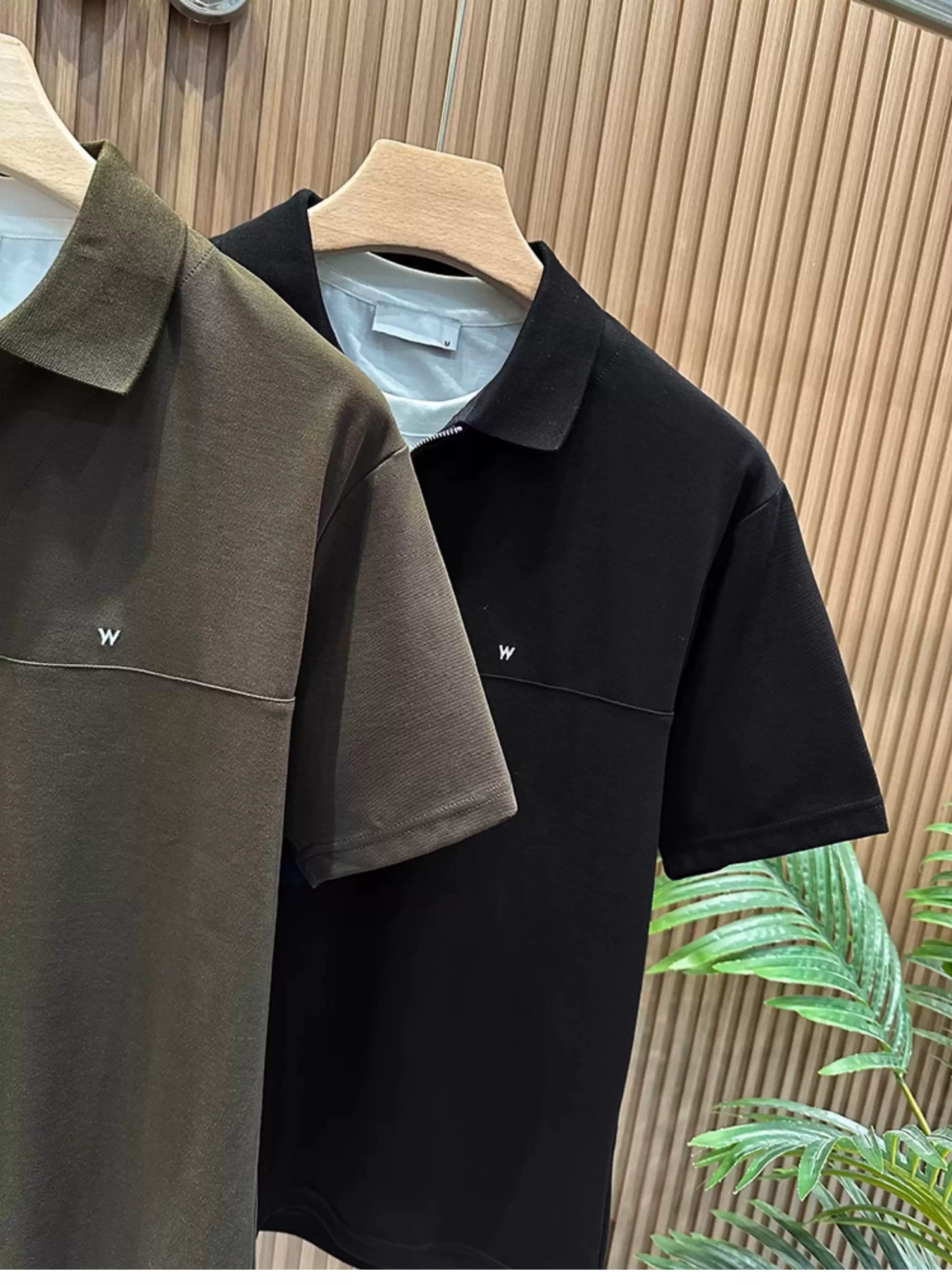 Front view of the Common Theory Signature Quarter-Zip Polo in black hanging on a wooden hanger, highlighting its ribbed collar, silver zipper, and structured silhouette. A dark olive variant is visible hanging directly behind it against a fluted wood wall paneling background.