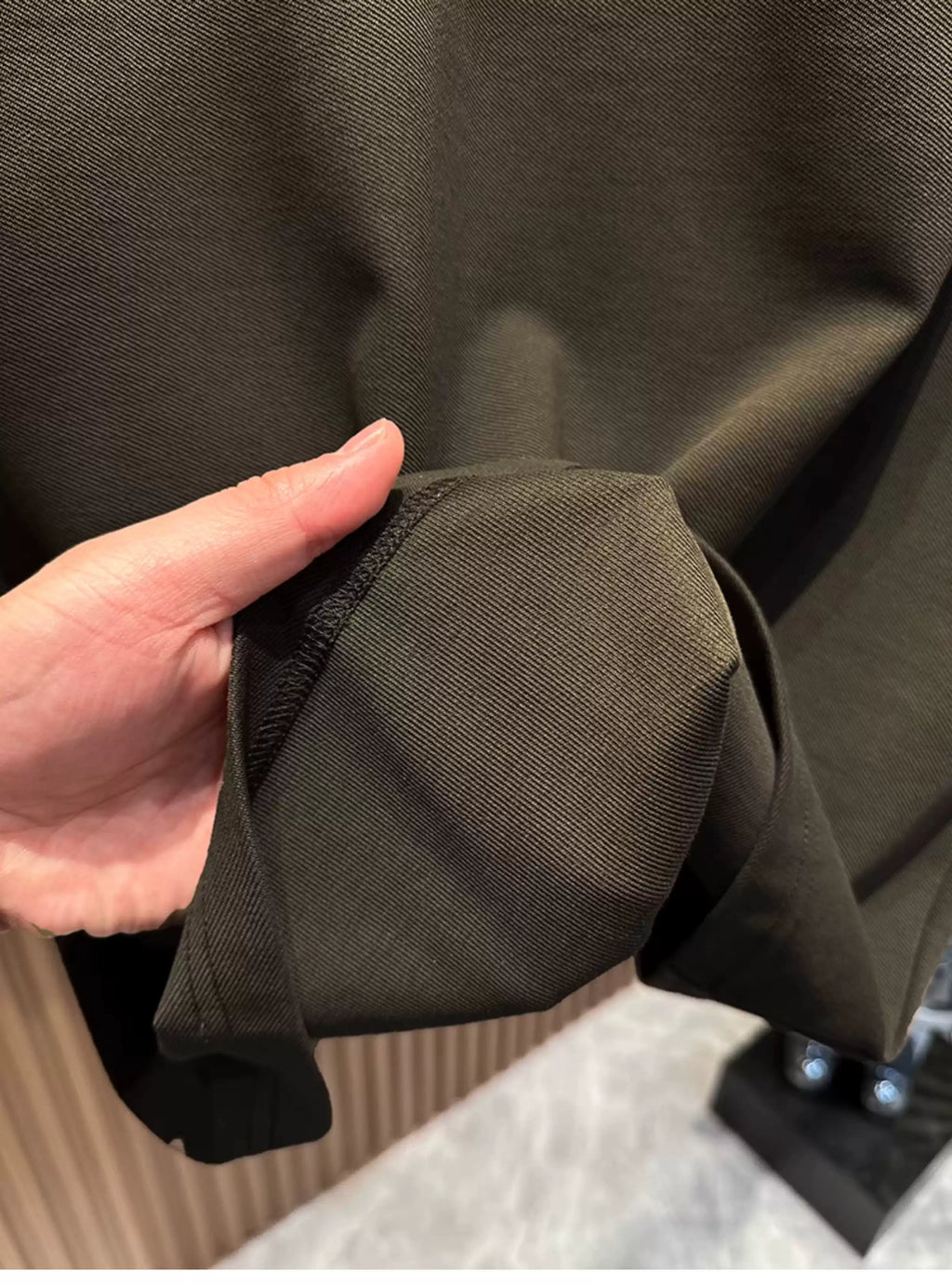 A close-up view of a person's hand gathering and holding the hem of the Common Theory Signature Quarter-Zip Polo in dark olive, highlighting its soft stretch heavyweight knit texture and clean, stitched edge.