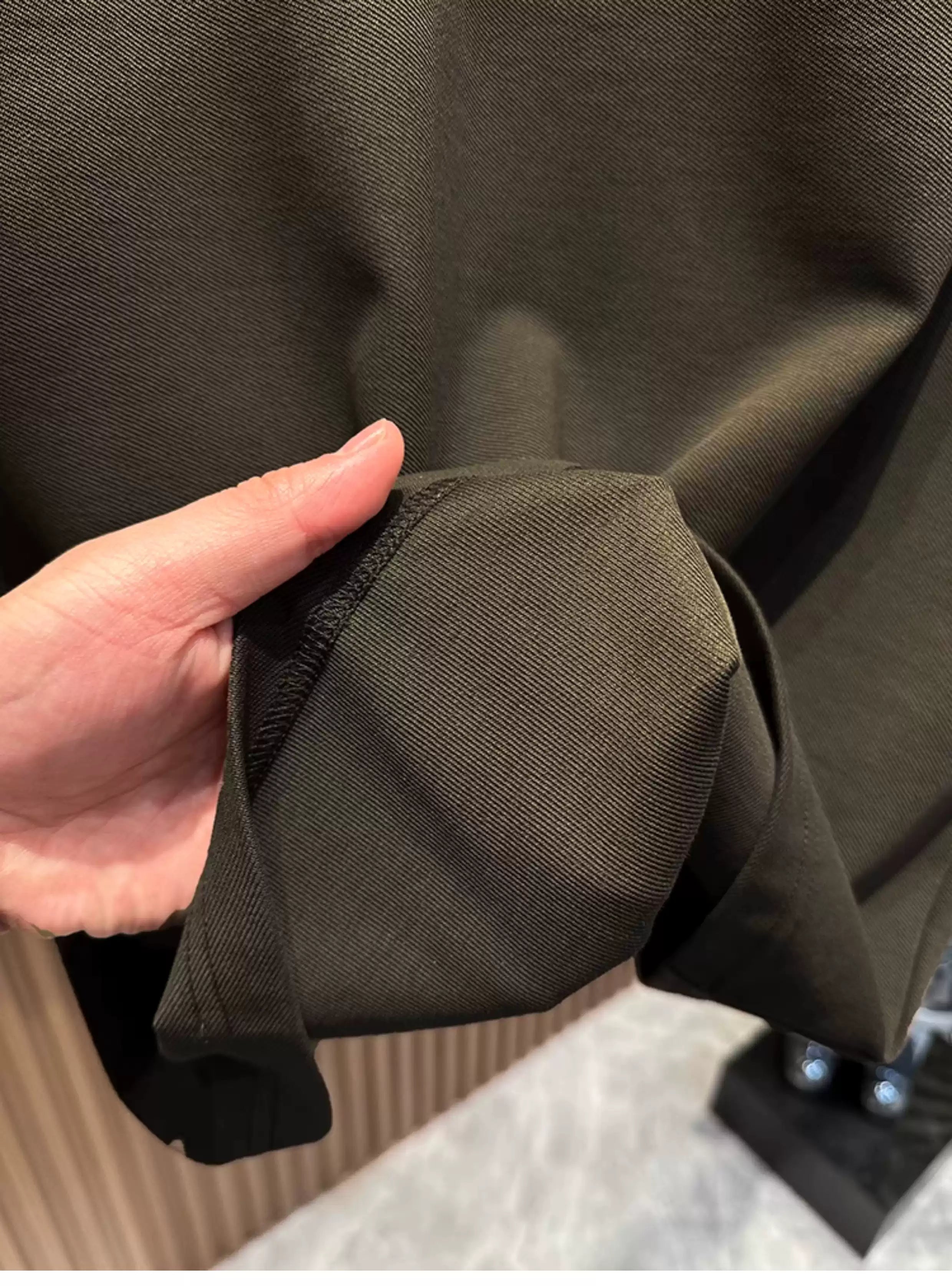 A close-up view of a person's hand gathering and holding the hem of the Common Theory Signature Quarter-Zip Polo in dark olive, highlighting its soft stretch heavyweight knit texture and clean, stitched edge.