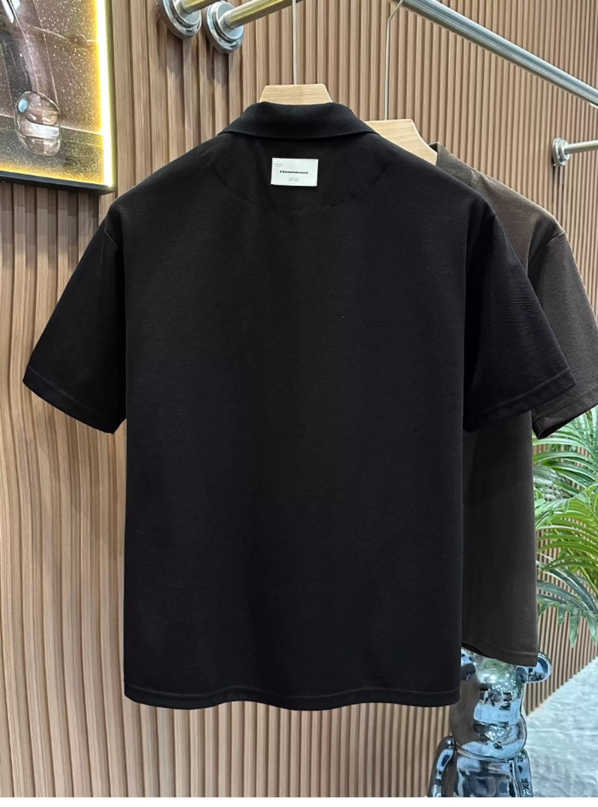 Rear view of the Common Theory Signature Quarter-Zip Polo in black hanging on a wooden hanger, displaying its clean finish, relaxed shoulder construction, and classic ribbed collar with a small white minimalist branding label underneath, against a fluted wood wall paneling background.