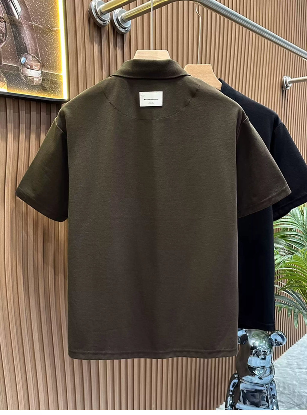 Rear view of the Common Theory Signature Quarter-Zip Polo in black hanging on a wooden hanger, highlighting its clean stitching, relaxed fit, and classic ribbed collar with a small white minimalist branding label underneath. A dark olive variant is visible hanging directly behind it against a fluted wood wall paneling background.