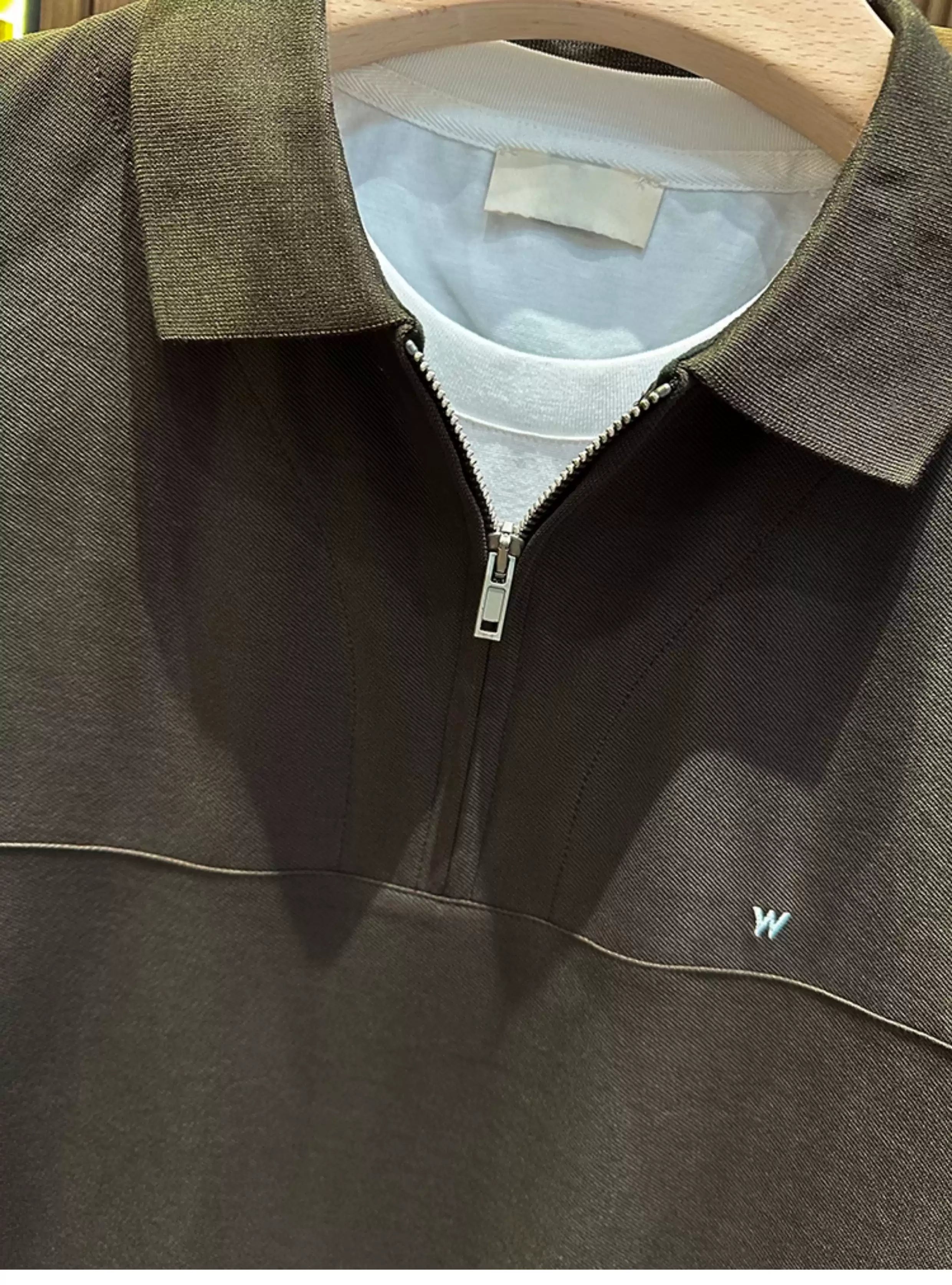 A close-up front view of the Common Theory Signature Quarter-Zip Polo in dark olive hanging on a wooden hanger, layered over a crisp white crewneck t-shirt. The image highlights the detailed piqué knit texture of the ribbed collar, the silver quarter-zip closure slightly unzipped, a clean mid-chest horizontal seam, and a small minimalist "W" or wave embroidered logo on the left chest.
