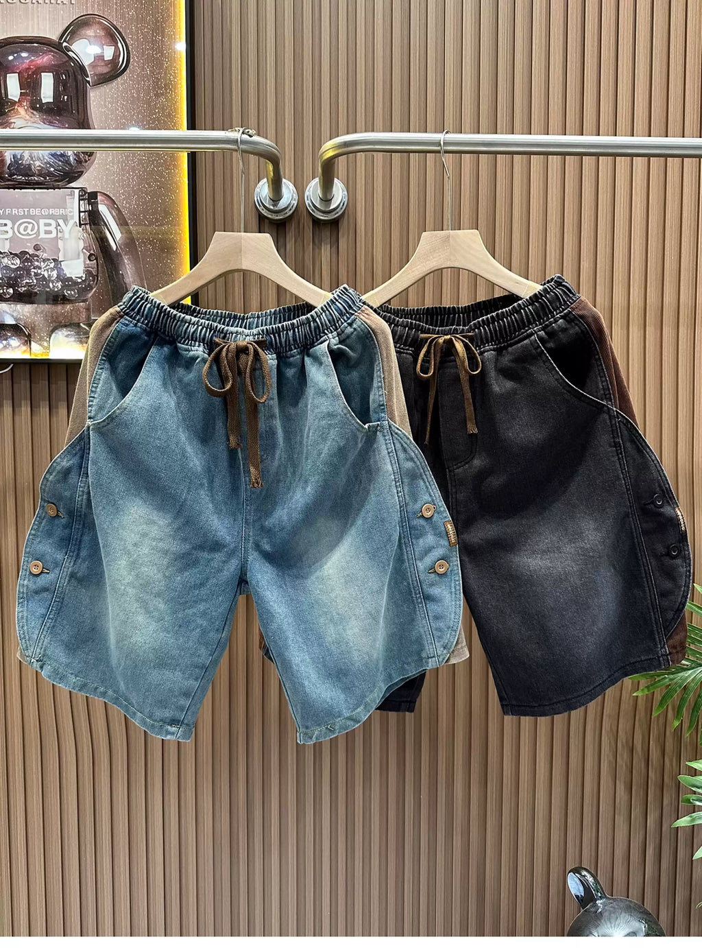 Front view of the Common Theory Vintage Panel Denim Shorts in blue and black hanging on a clothing rack, highlighting their relaxed oversized fit, elastic waistband with drawstrings, and unique paneled side button detailing.
