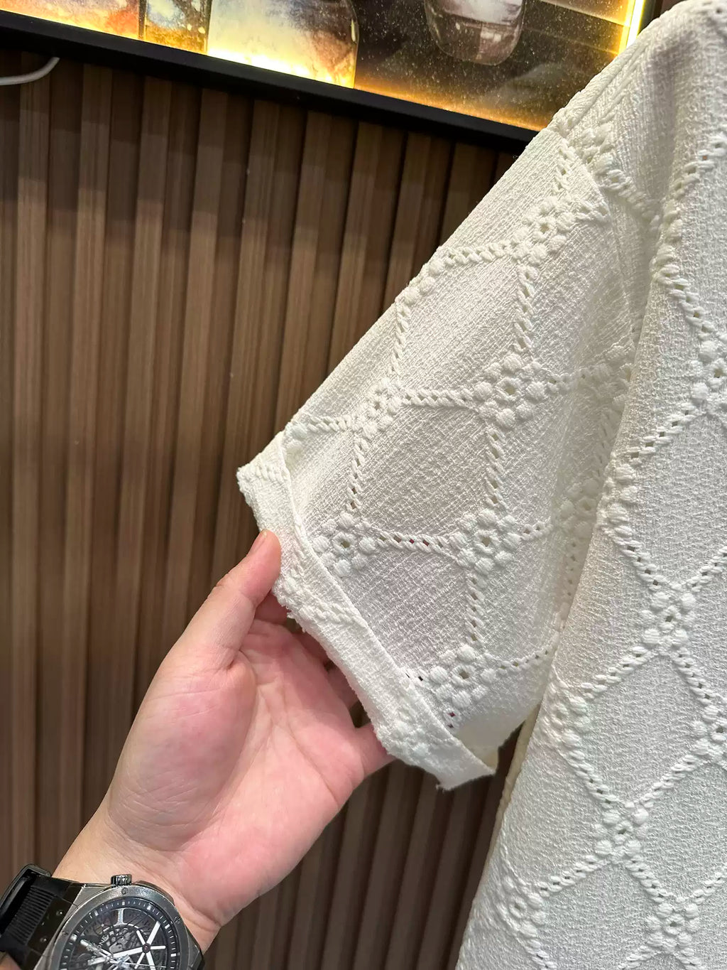 Close-up of a person's hand holding up the short sleeve of an ivory white textured knit shirt, showcasing the intricate diamond and floral open-knit pattern against a dark wood-paneled wall backdrop.