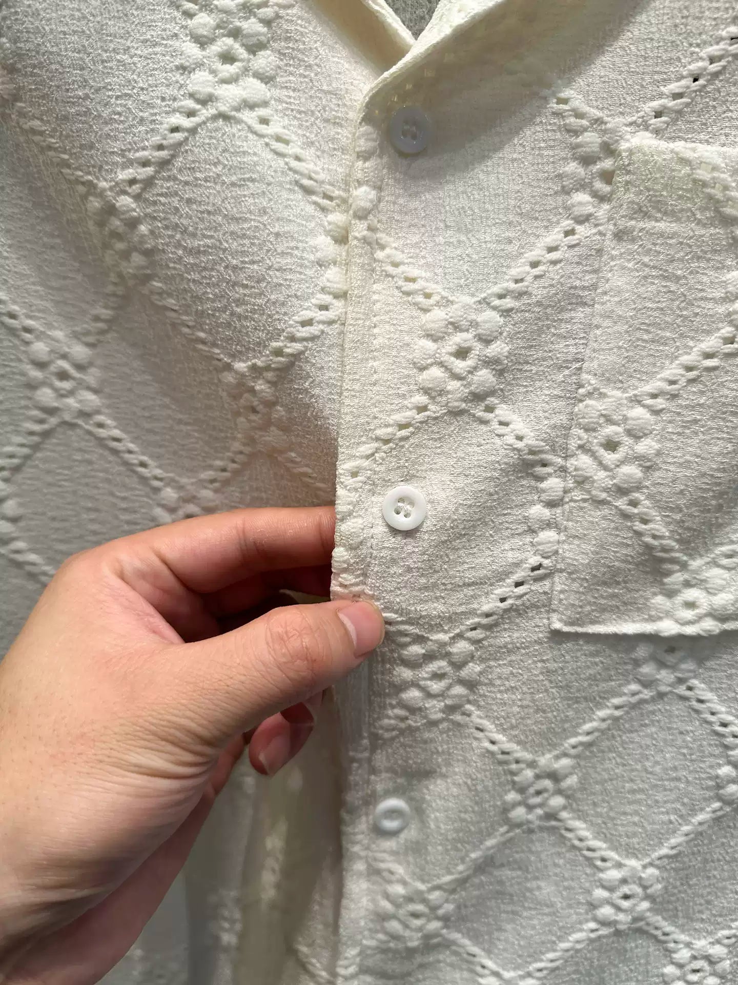 Close-up of the Common Theory Riviera Ivory White Resort Shirt, showing a person's hand touching the front placket next to the white buttons, highlighting the intricate diamond and floral open-knit pattern and the chest pocket.
