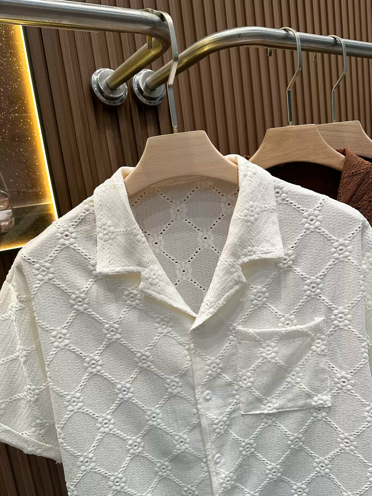 Close-up of the Common Theory Riviera Ivory White Resort Shirt on a wooden hanger, showcasing its relaxed camp collar, chest pocket, and the intricate open-knit diamond and floral pattern against a fluted wood wall paneling background.