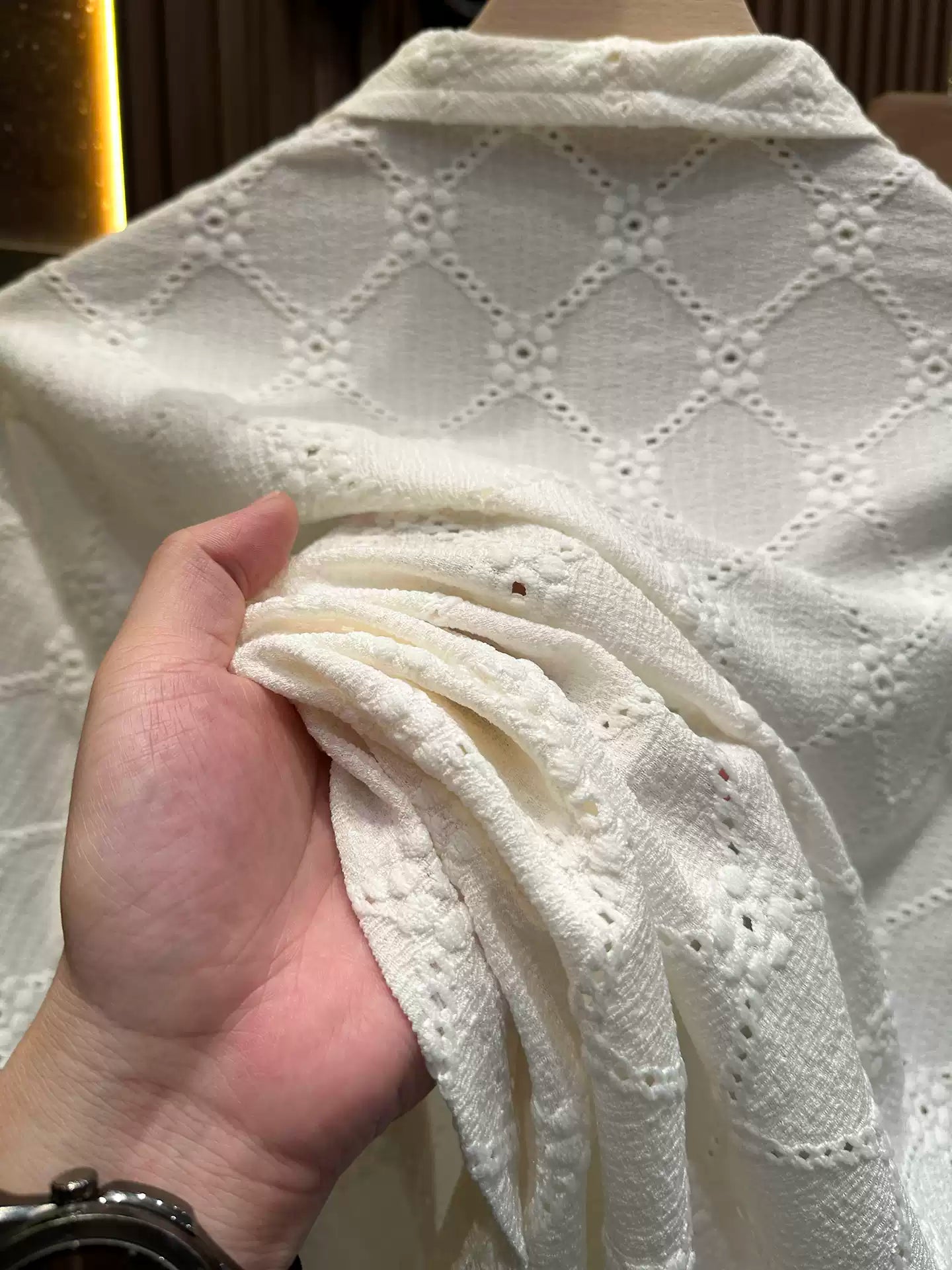 A close-up view of a person's hand holding up a section of the Common Theory Riviera Ivory White Resort Shirt, highlighting the soft fabric texture and the detailed geometric diamond and floral open-knit pattern