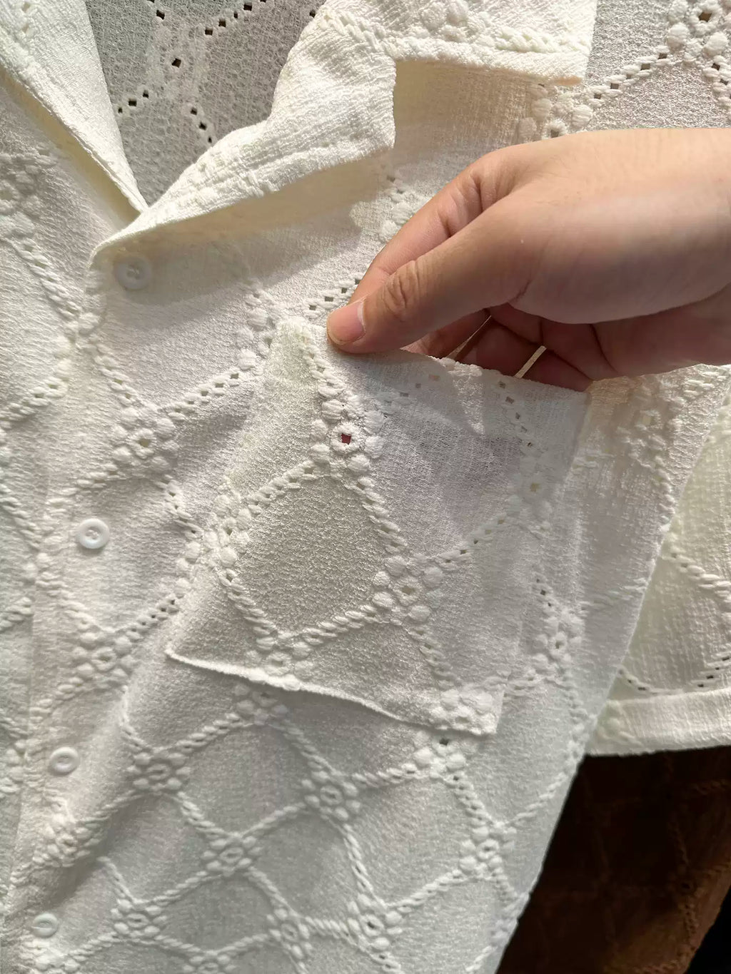 Close-up of the Common Theory Riviera Ivory White Resort Shirt, showing a person's hand pulling open the front chest pocket, highlighting the intricate geometric diamond and floral open-knit pattern and the short sleeve.