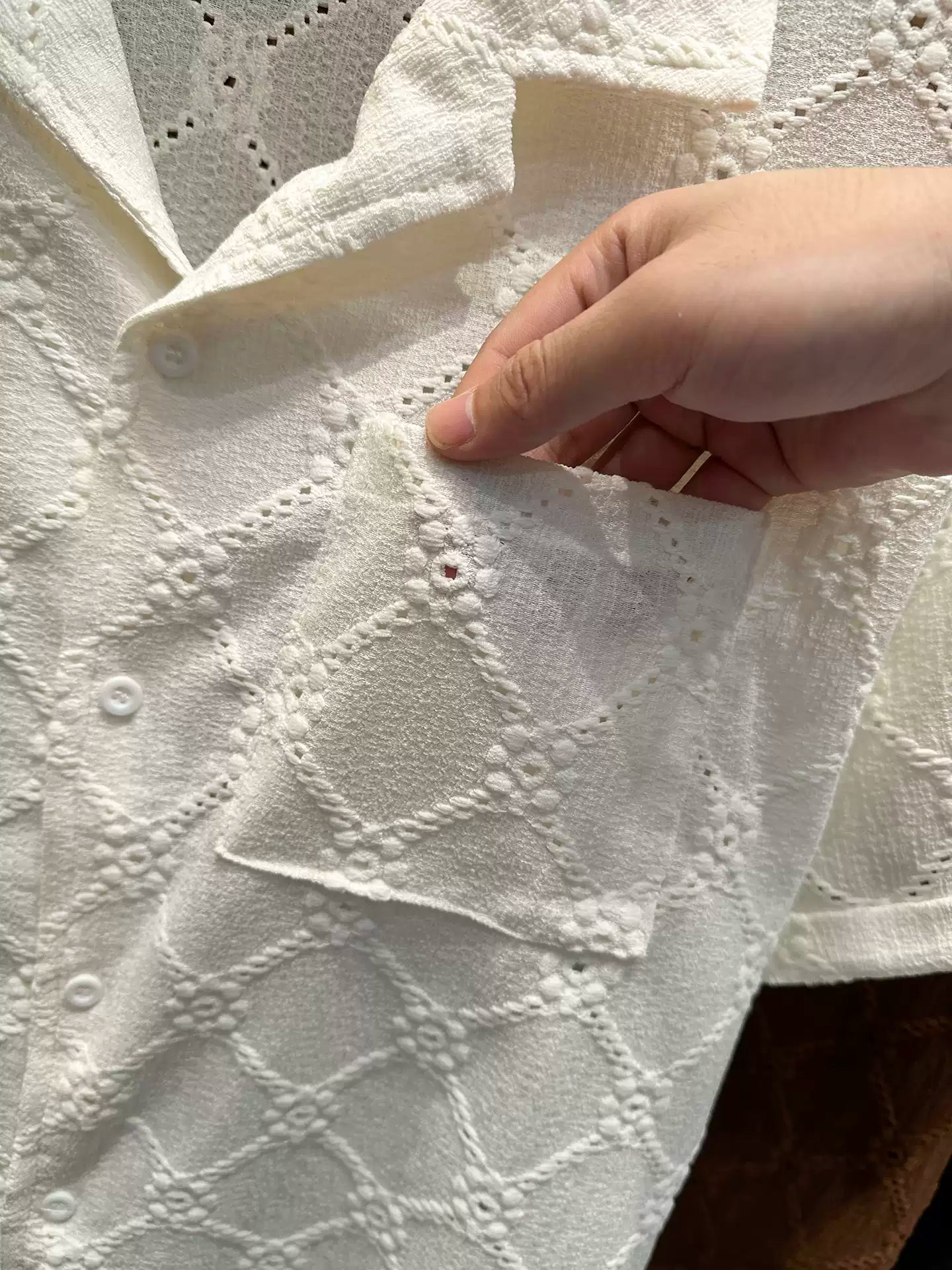 Close-up of the Common Theory Riviera Ivory White Resort Shirt, showing a person's hand pulling open the front chest pocket, highlighting the intricate geometric diamond and floral open-knit pattern and the short sleeve.