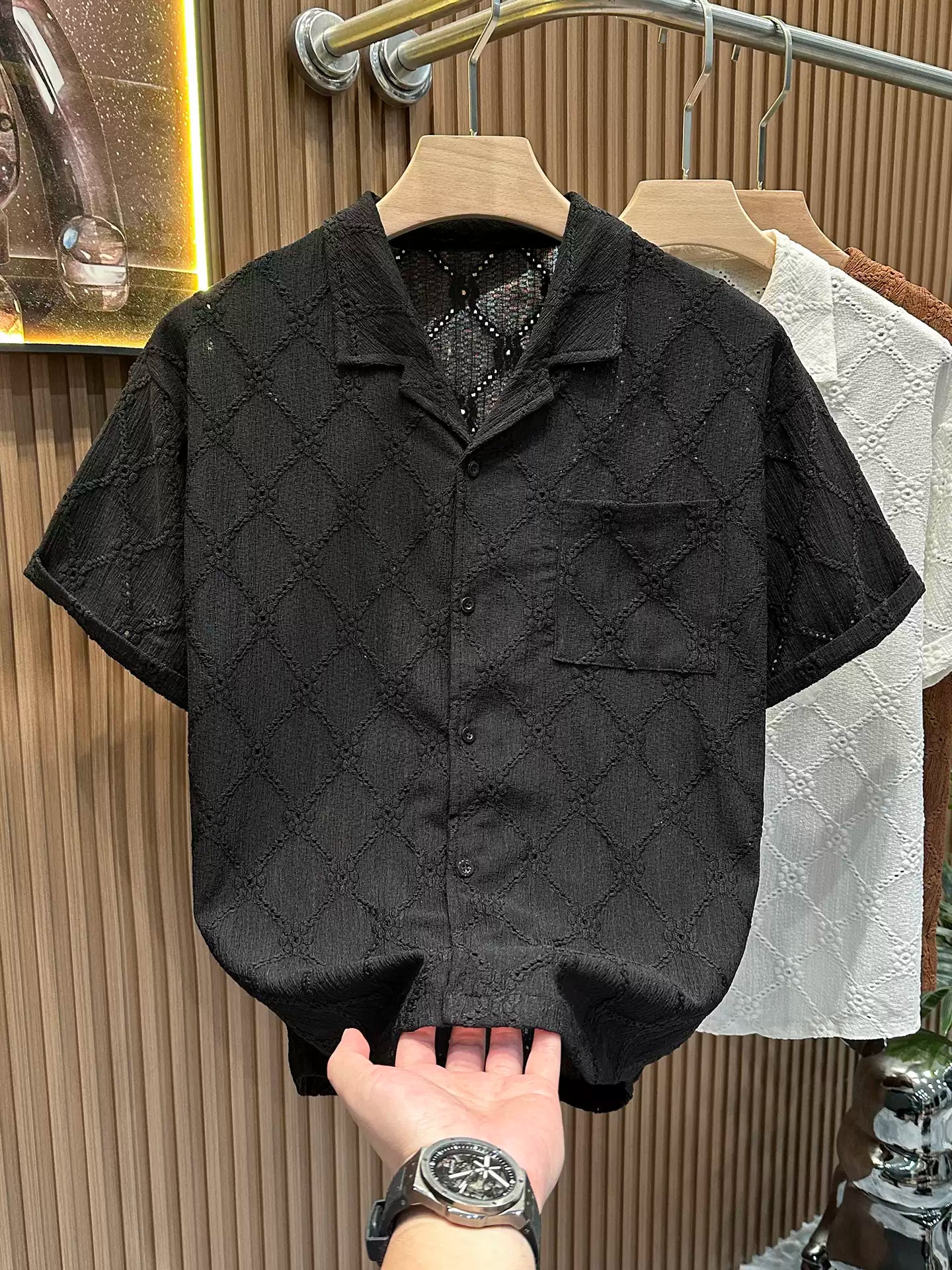 Close-up of the Common Theory Riviera Midnight Black Resort Shirt on a wooden hanger, featuring a distinctive geometric diamond and floral open-knit pattern, a relaxed camp collar, and a front chest pocket, with a hand touching the bottom hem.