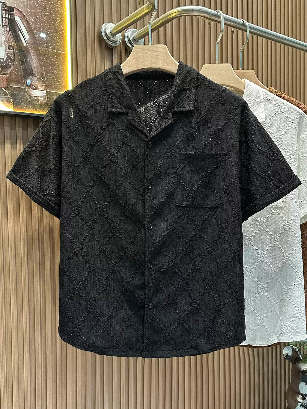 The Common Theory Riviera Midnight Black Resort Shirt hanging on a wooden clothes hanger, showcasing its relaxed camp collar, chest pocket, and intricate geometric diamond open-knit pattern, with the ivory white and cocoa brown variants visible hanging behind it